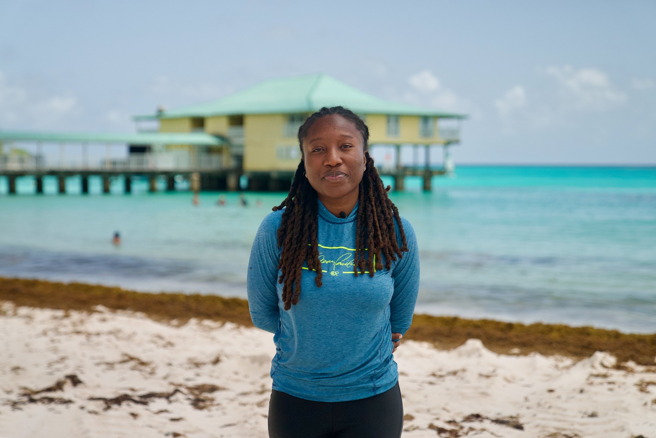 Bióloga marinha Micaela Small em Brownes Beach, com uma doca no fundo e o sargasso dividindo a areia da água cristalina