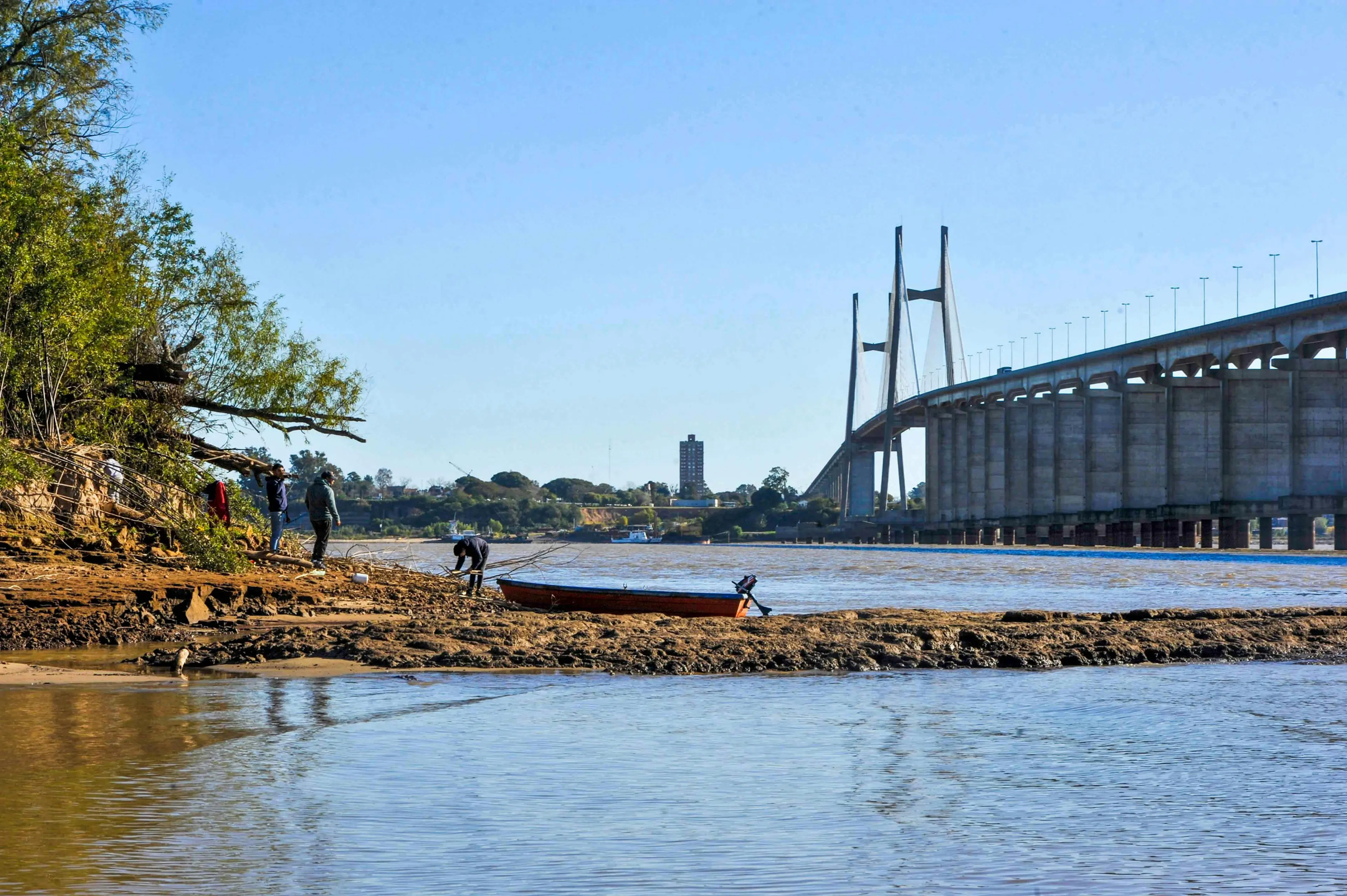 A boat rests on the riverban with a bridge in the background 