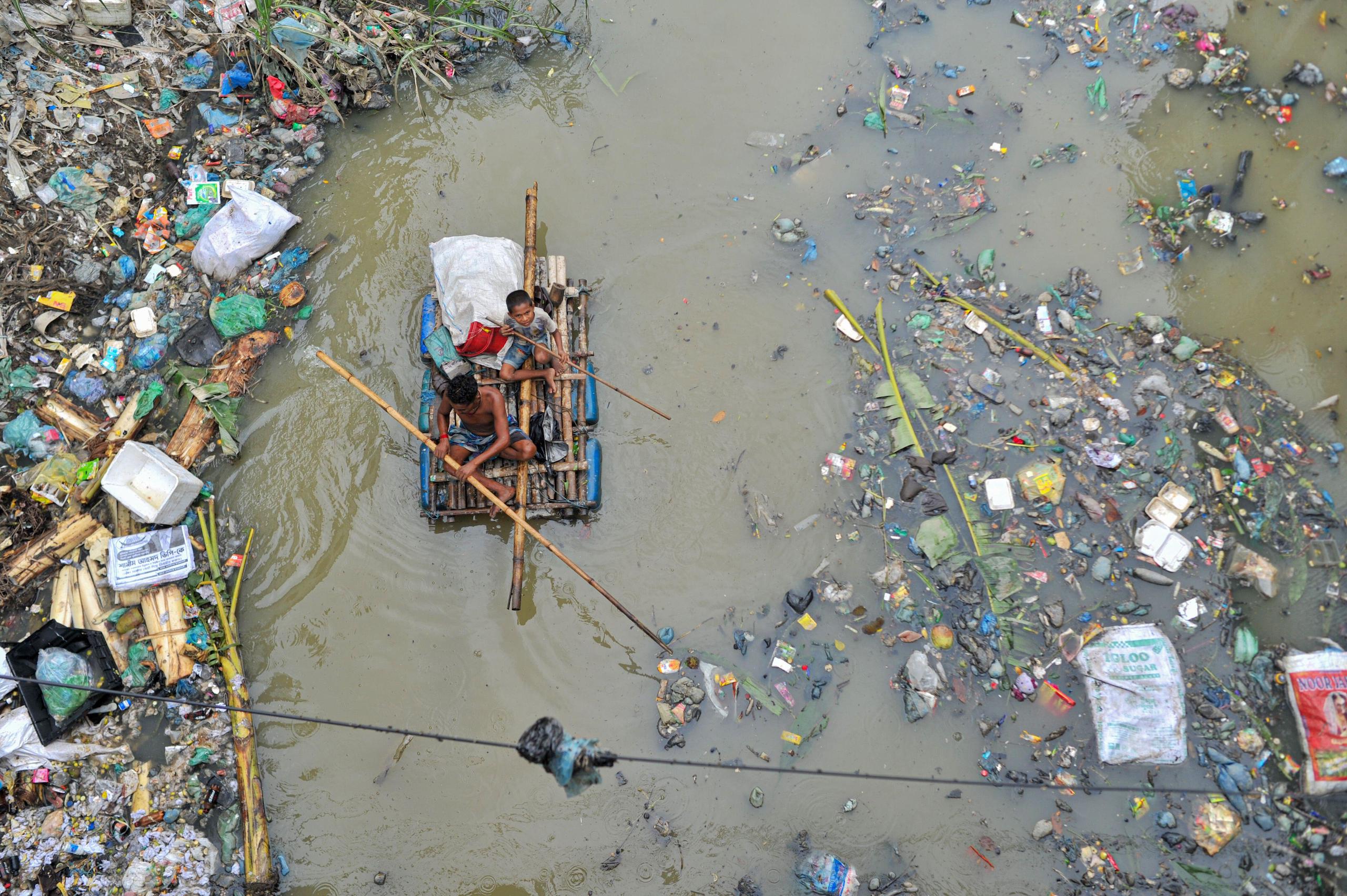 <p>A father and son rowing on a handmade raft searching for plastic waste to recycle in a canal of the Kazir Bazar area of Sylhet, Bangladesh. This canal is directly connected with the Surma River and is polluting the river and its river bed. Huge piles of polythene are obstructing navigation and causing scarcity of usable water from the Surma River of Bangladesh. (Image: Rafayat Khan / Majority World CIC / Alamy)</p>