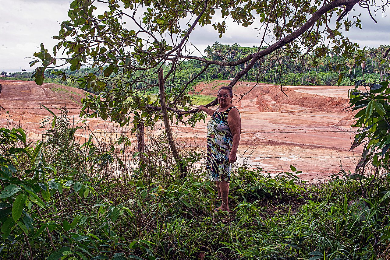 Moradora de meia-idade, com vestido colorido e chinelos, posa em meio à vegetação, com solo arenoso no fundo