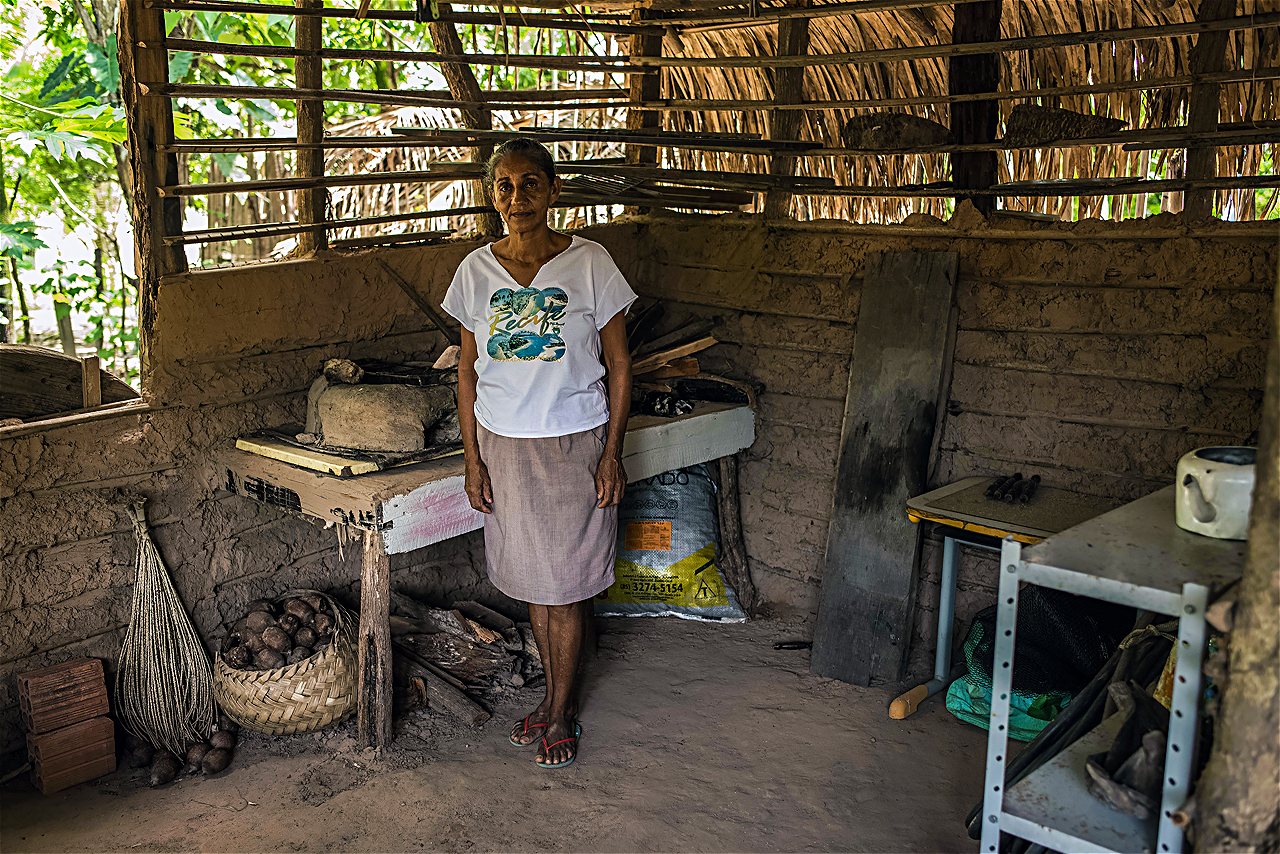 Mulher com camiseta branca e saia na cozinha de uma casa de barro, com fogão à lenha no fundo