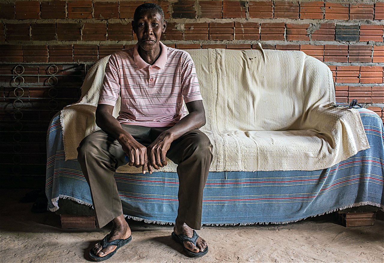 homem negro idoso, alto, de calça escura, chinelo e camisa listrada, sentado em sofá apoiado por tijolos, em casa com tijolos à mostra