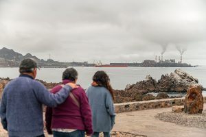 tres personas de espaldas, mirando hacia torres humeantes en la costa