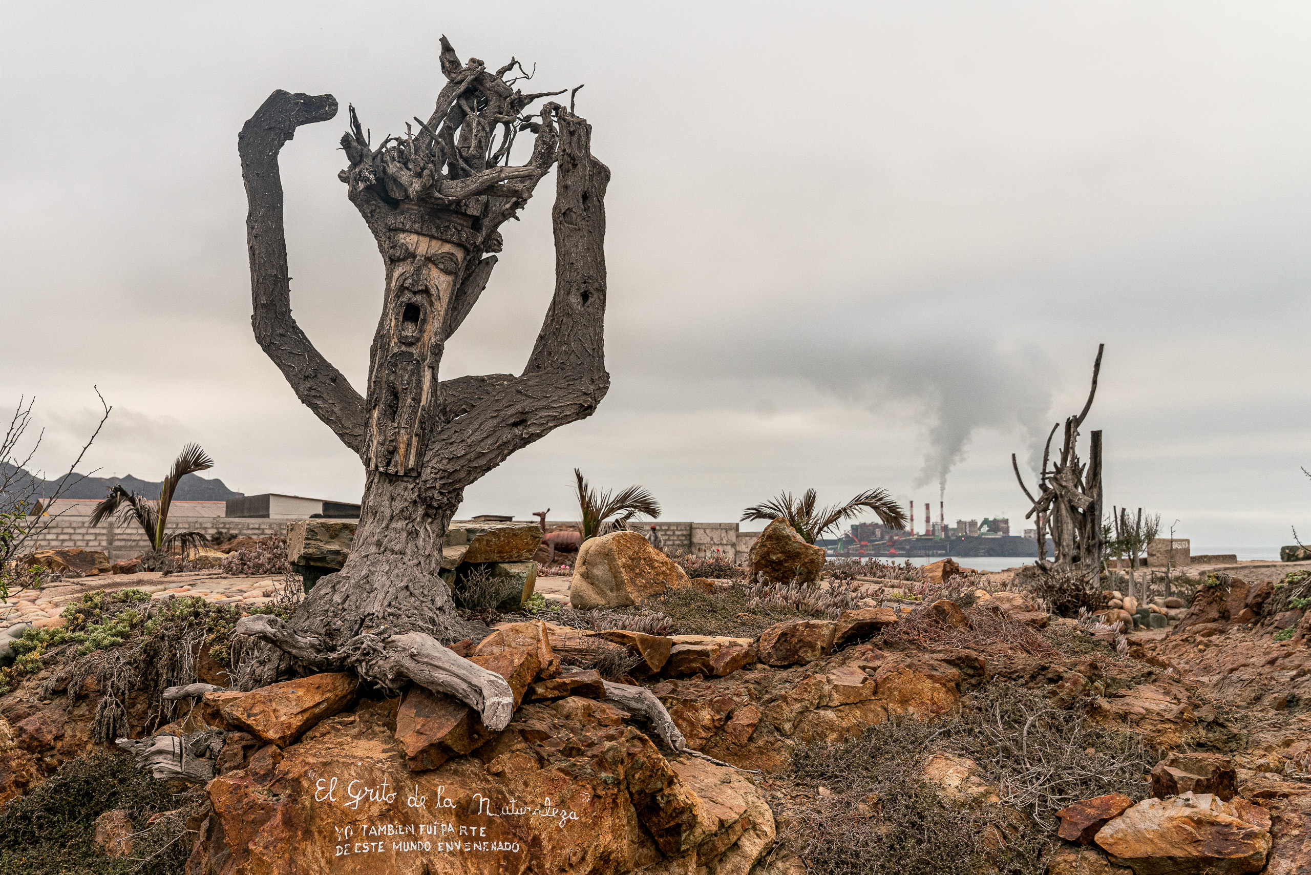 Escultura de Luis Alberto Triviño perto da costa de Huasco, com a zona industrial ao fundo. A obra é identificada na parte de baixo sob o título O grito da natureza: Também fiz parte deste mundo envenenado.