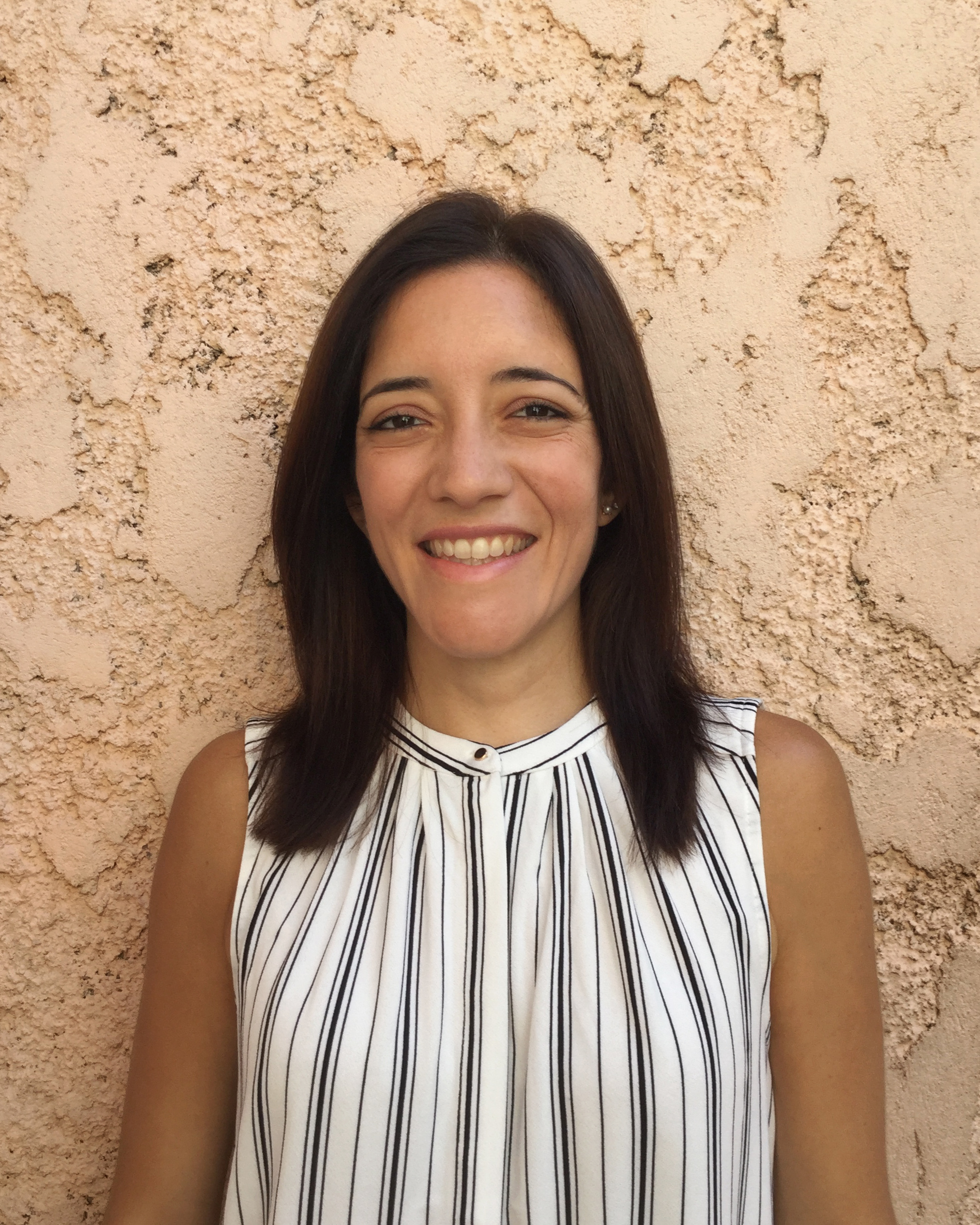 retrato de Juliana González Jáuregui. Mulher de pele morena clara, com cabelo castanho liso e comprimento até os ombros. Elaestá sorrindo e uma camisa social listrada, sem mangas.