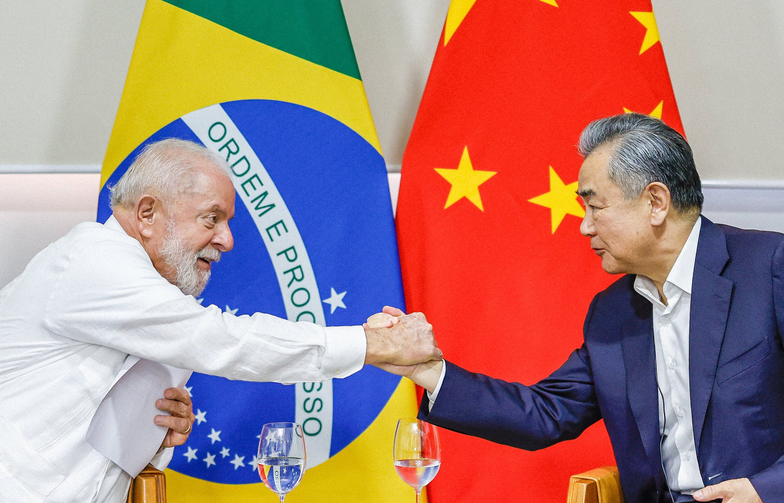 Lula aperta a mão do chanceler chinês Wang Yi, com a bandeira do Brasil e da China ao fundo
