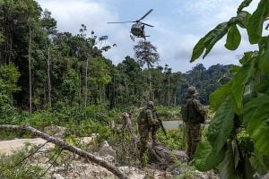 A helicopter hovers above a forest, with soldiers stationed below