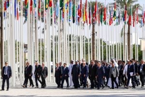 group of people in suits walking beneath limp flags on tall poles