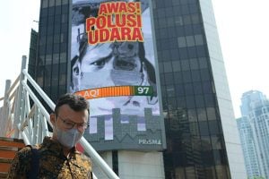 A man wearing a mask walks on a bridge past a billboard that says "Beware of Air Pollution"