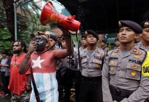 man with red, white and blue body paint speaking into megaphone and surrounded by other men in uniform