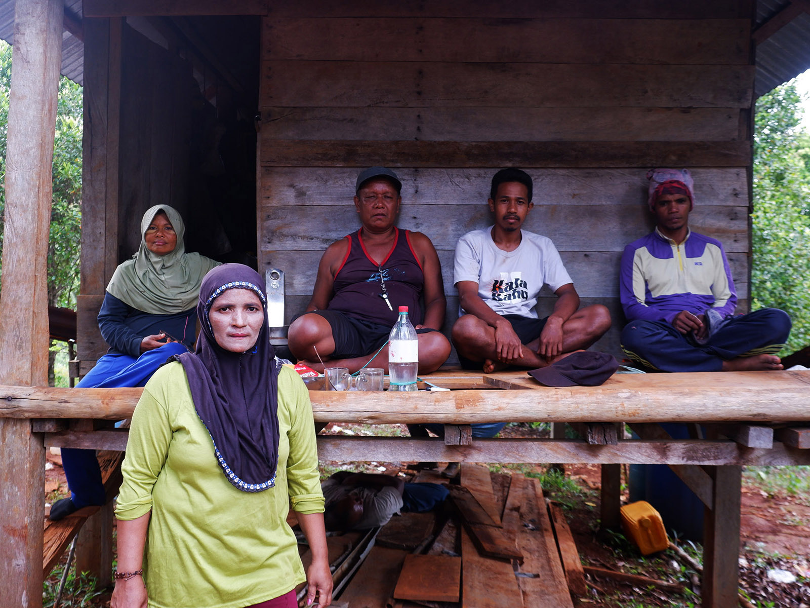 A woman in a hijab, stands in front of a shelter where four people sits there