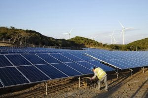 A worker maintains a solar panel array with wind turbines in the background.