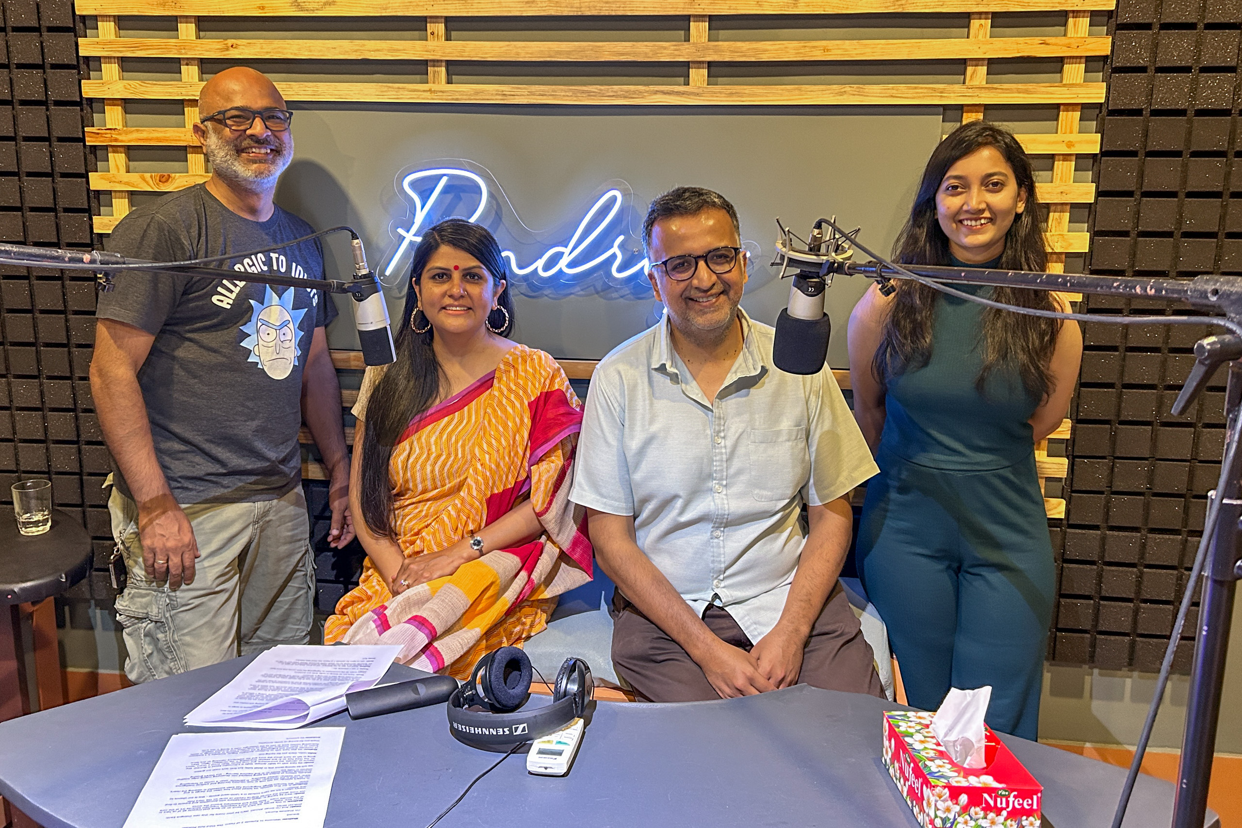 Four people are standing and sitting in a podcast studio with microphones and a neon sign in the background.