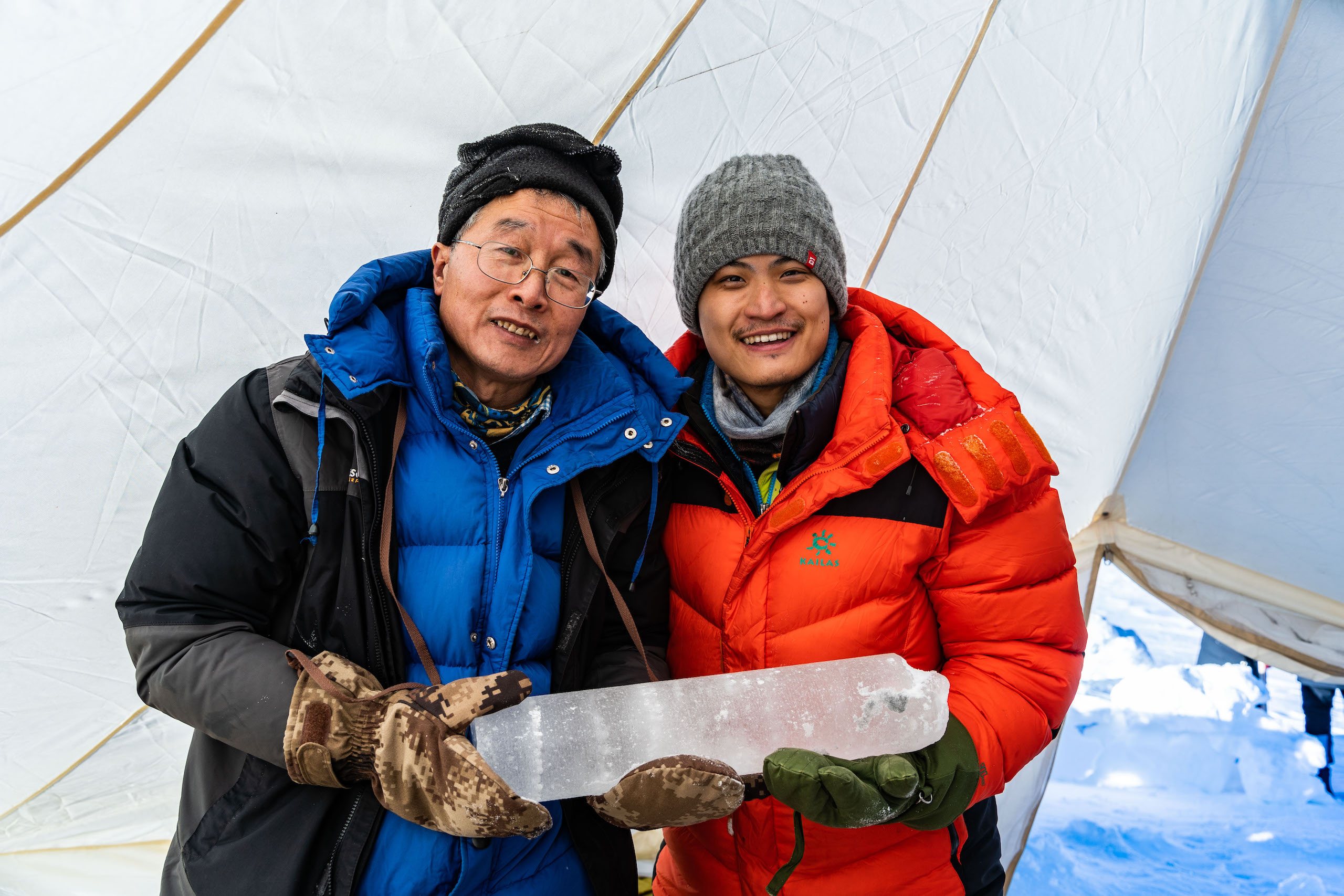Two men in winter gear are holding a large block of ice together to 