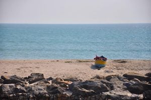 A colorful boat with gear on a sandy beach by the sea, behind a rocky barrier