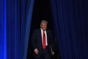 Donald Trump in a dark suit and red tie walks out from behind blue curtains, holding a piece of paper in his left hand.