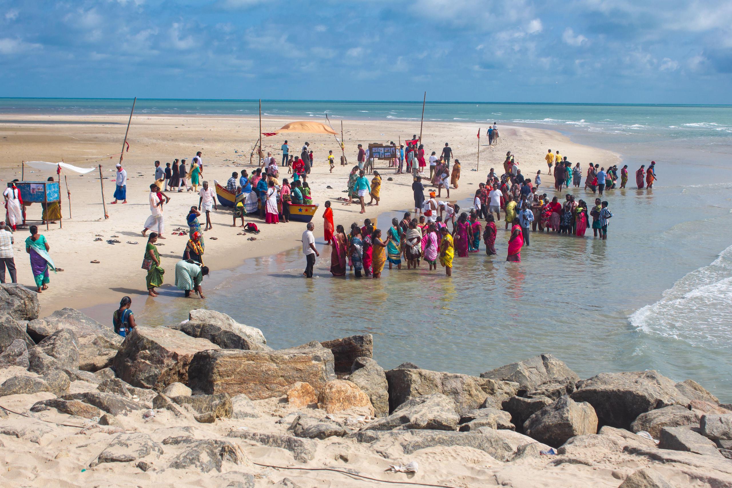 Crowd of people in colorful clothing on a sandy beach with waves and boats nearby.