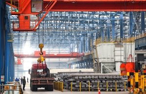 A crane in a vast warehouse is lifting and loading steel to a truck