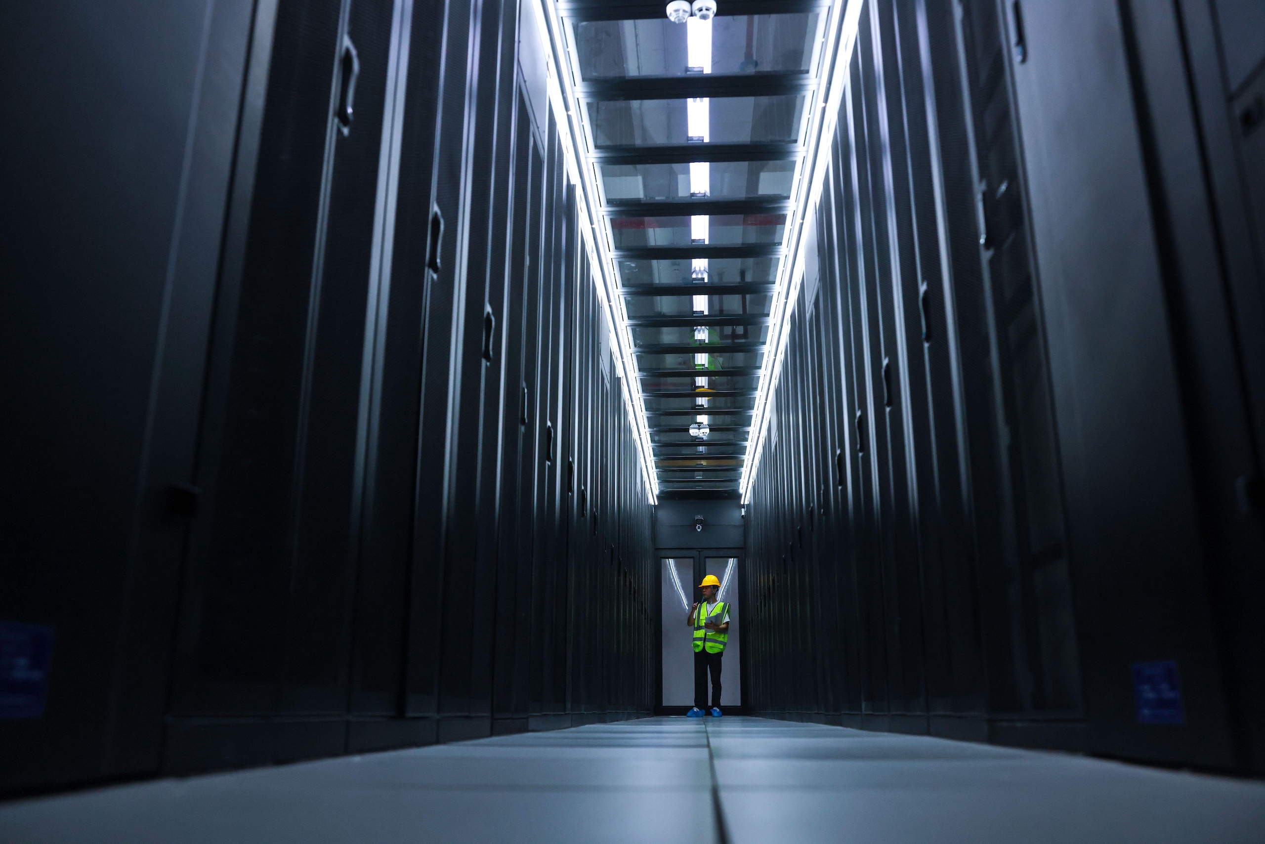 A person in a safety vest and helmet stands in a dimly lit data centre aisle 