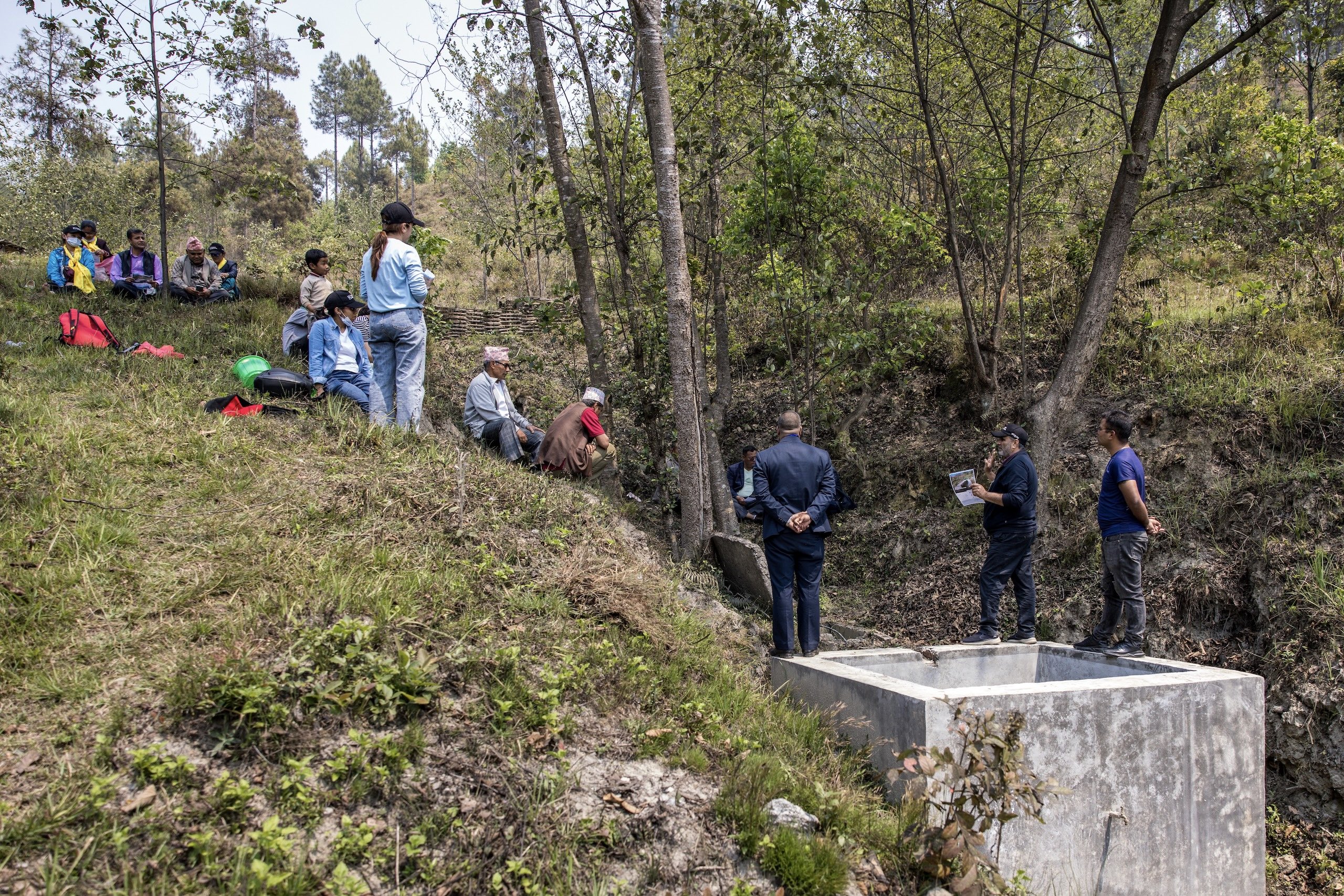 A group of people sits on a grassy slope while others stand by a concrete structure in a wooded area