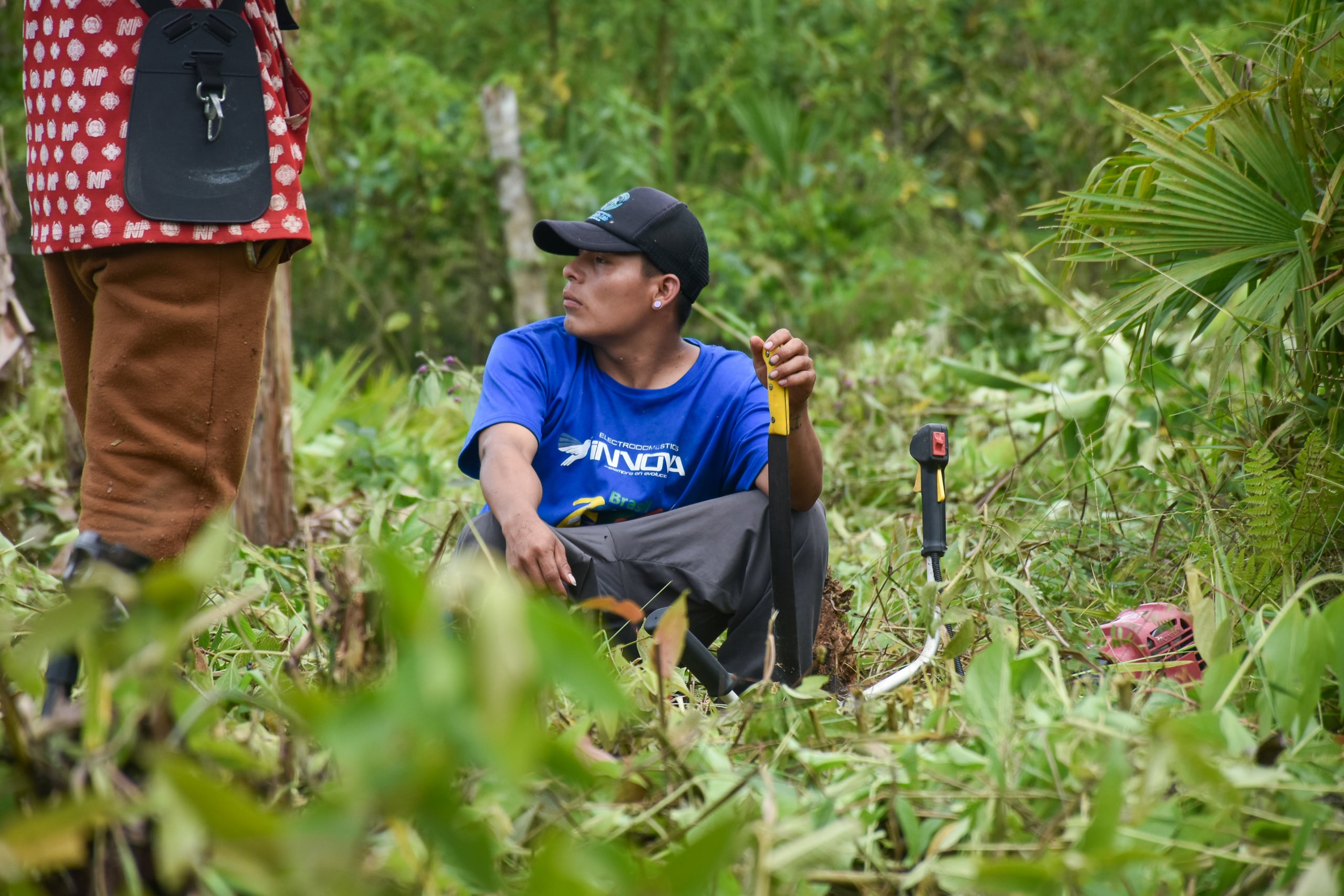 A man kneels in the grass, holding a gardening tool