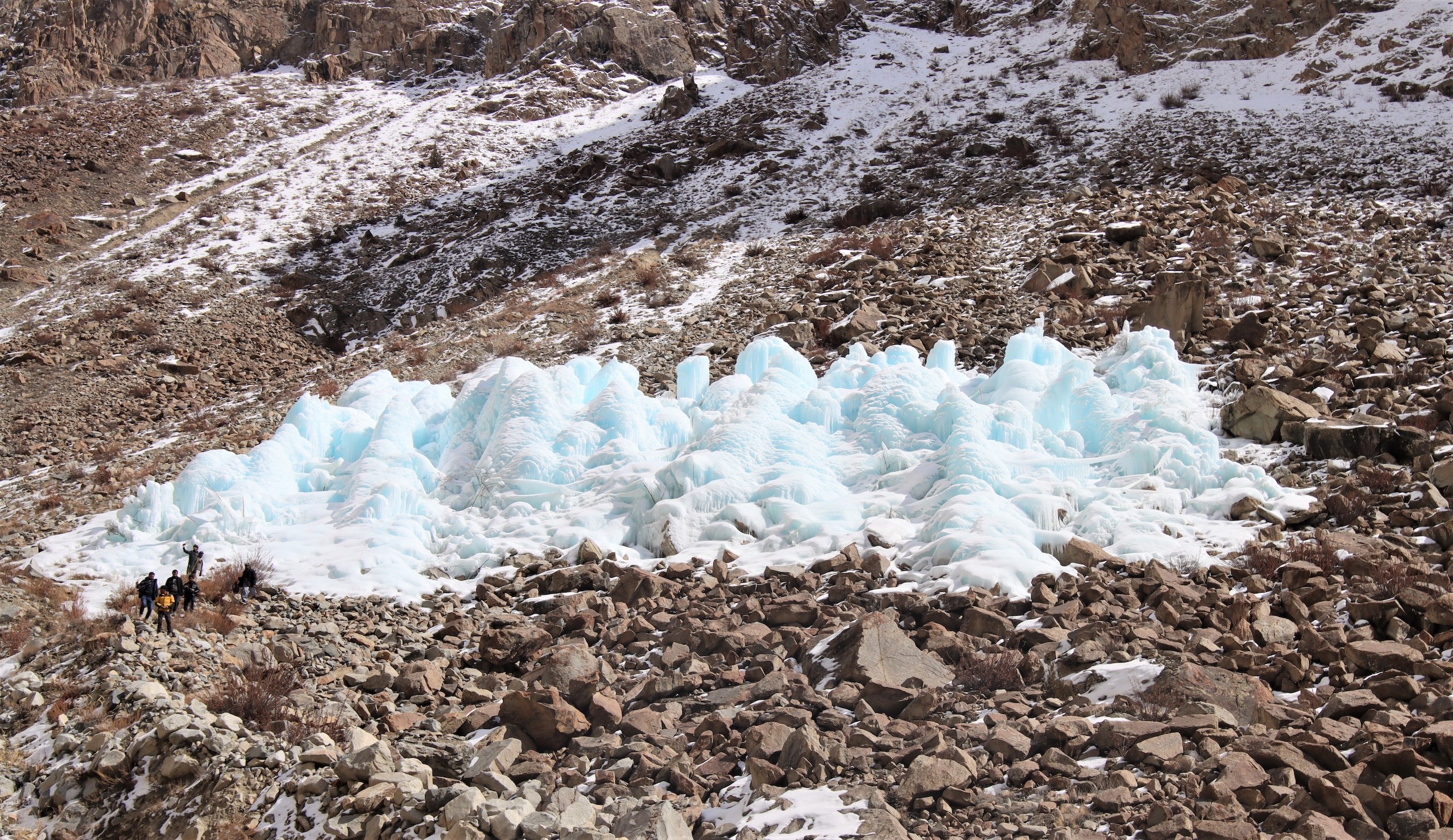 A group of people walk near a large ice stupas surrounded by rocky terrain and patches of snow in a mountainous area.