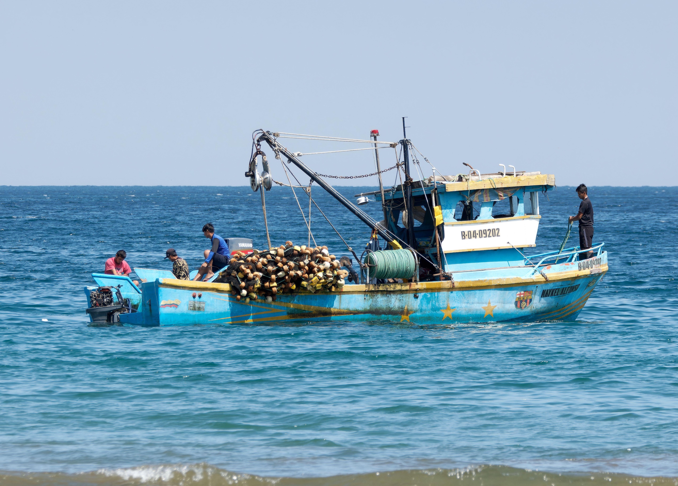 A fishing boat filled with traps and ropes, with several fishermen working aboard against a blue ocean backdrop.