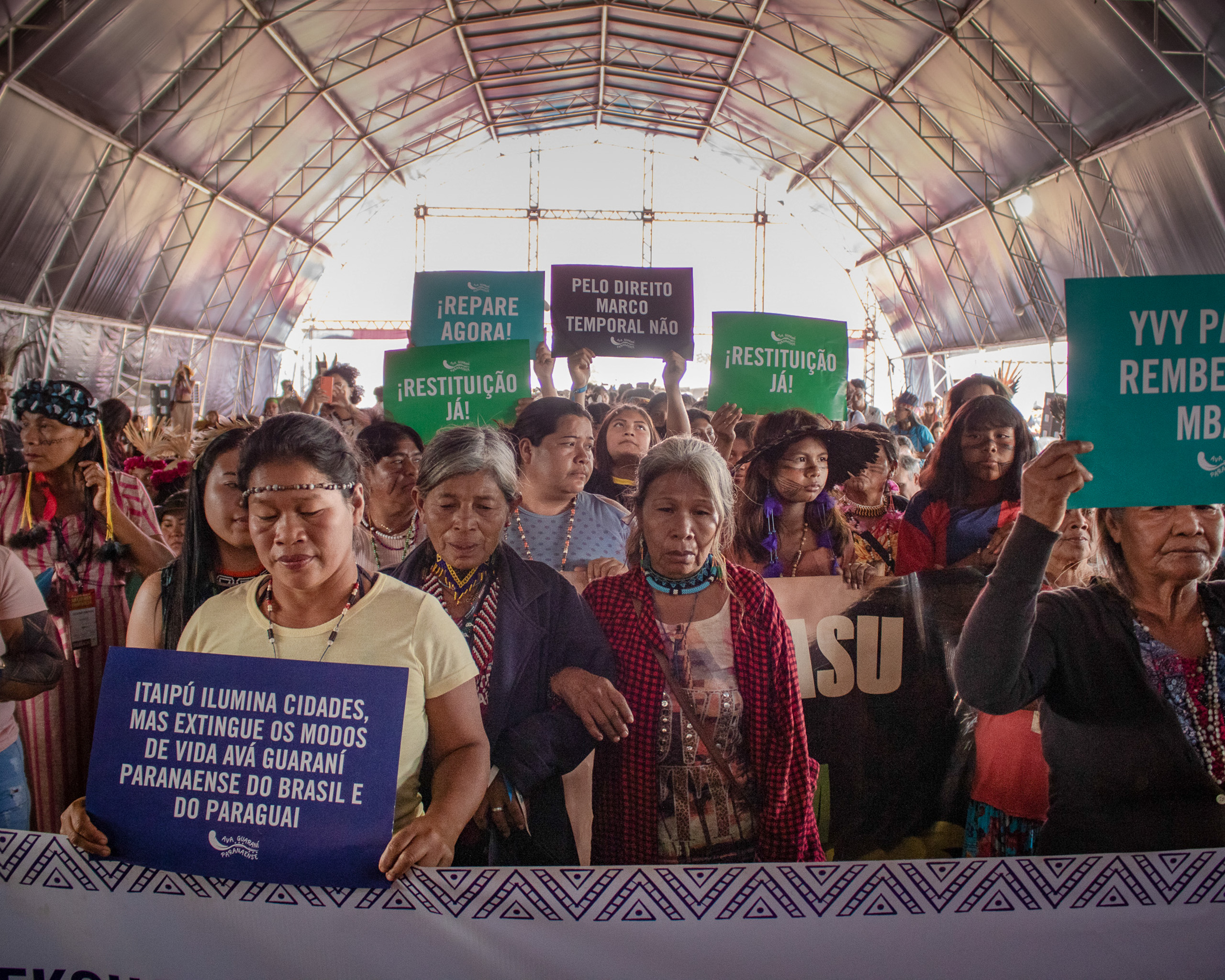 A diverse group of indigenous women inside a tent, holding signs with various messages