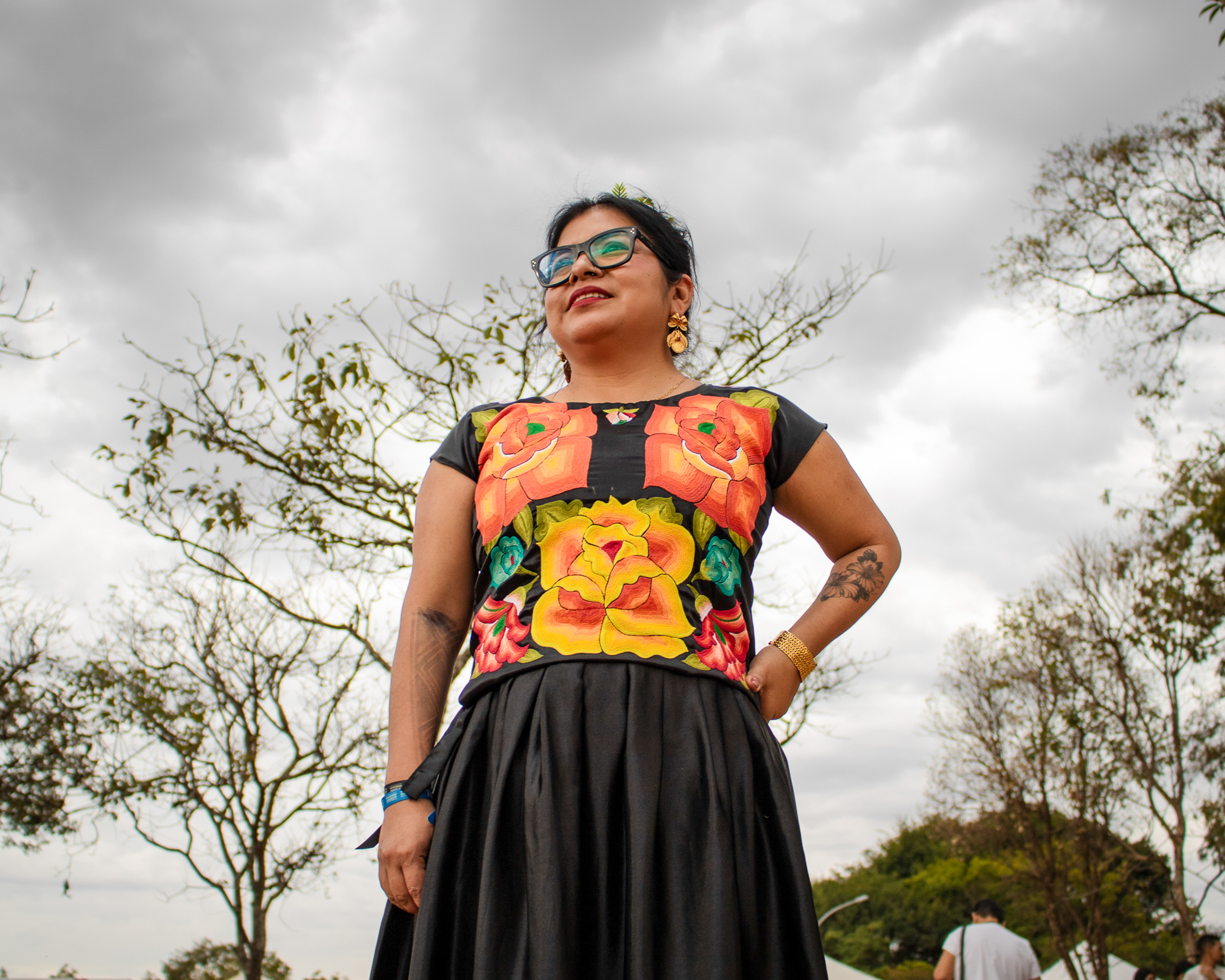 A woman stands confidently in a flower-patterned top and a black skirt, surrounded by leafless trees under a cloudy sky