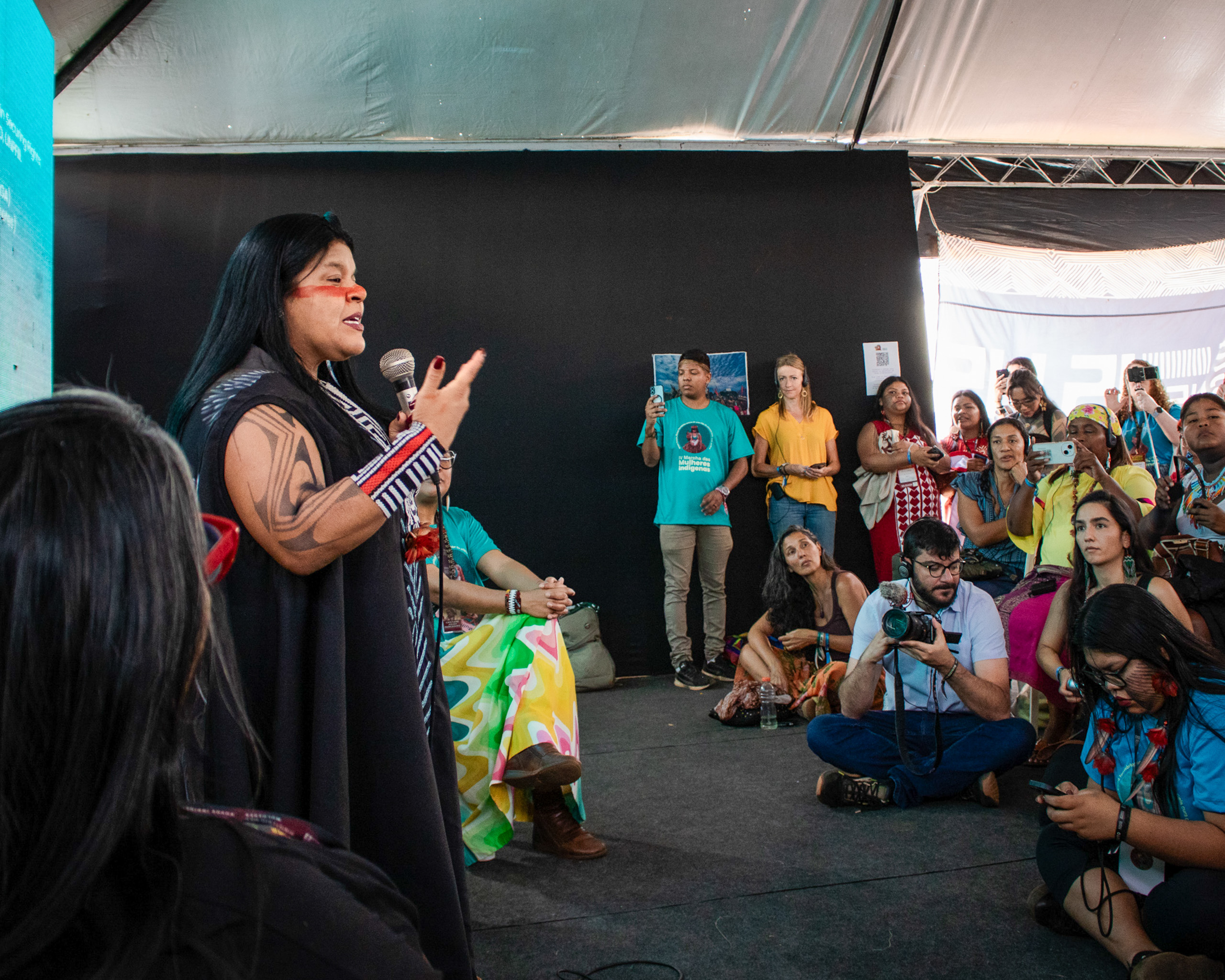 A female speaker engages an audience in a tent, while attendees capture the moment with their cameras and phones