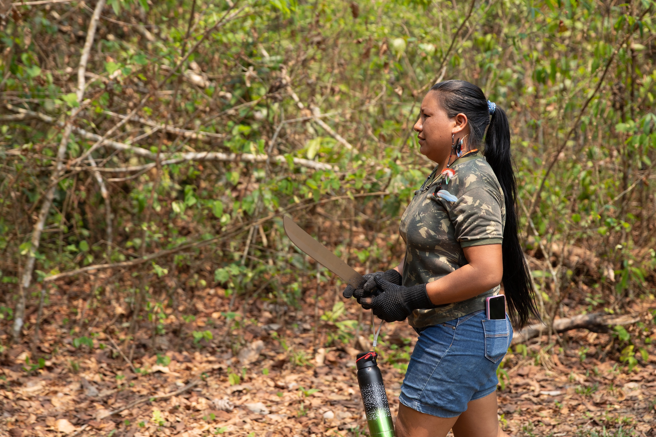 A woman stands in a lush green forest, holding a machete and wearing gloves