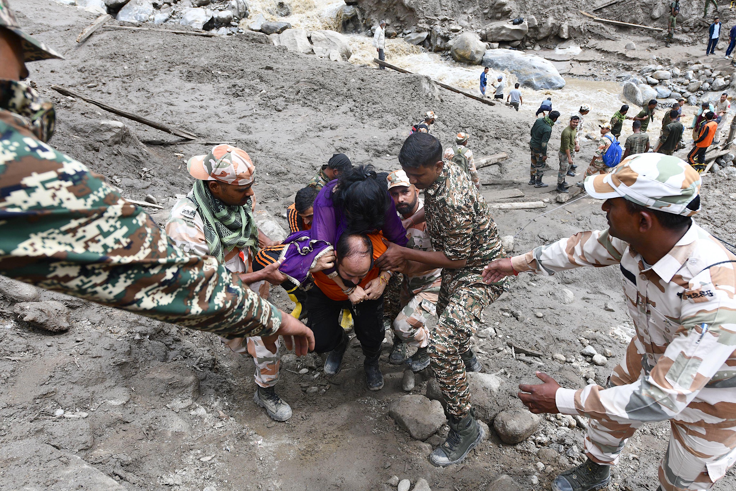 Rescue workers assist a person in distress, navigating rough terrain with rocks and a river in the background, during a flooding response.
