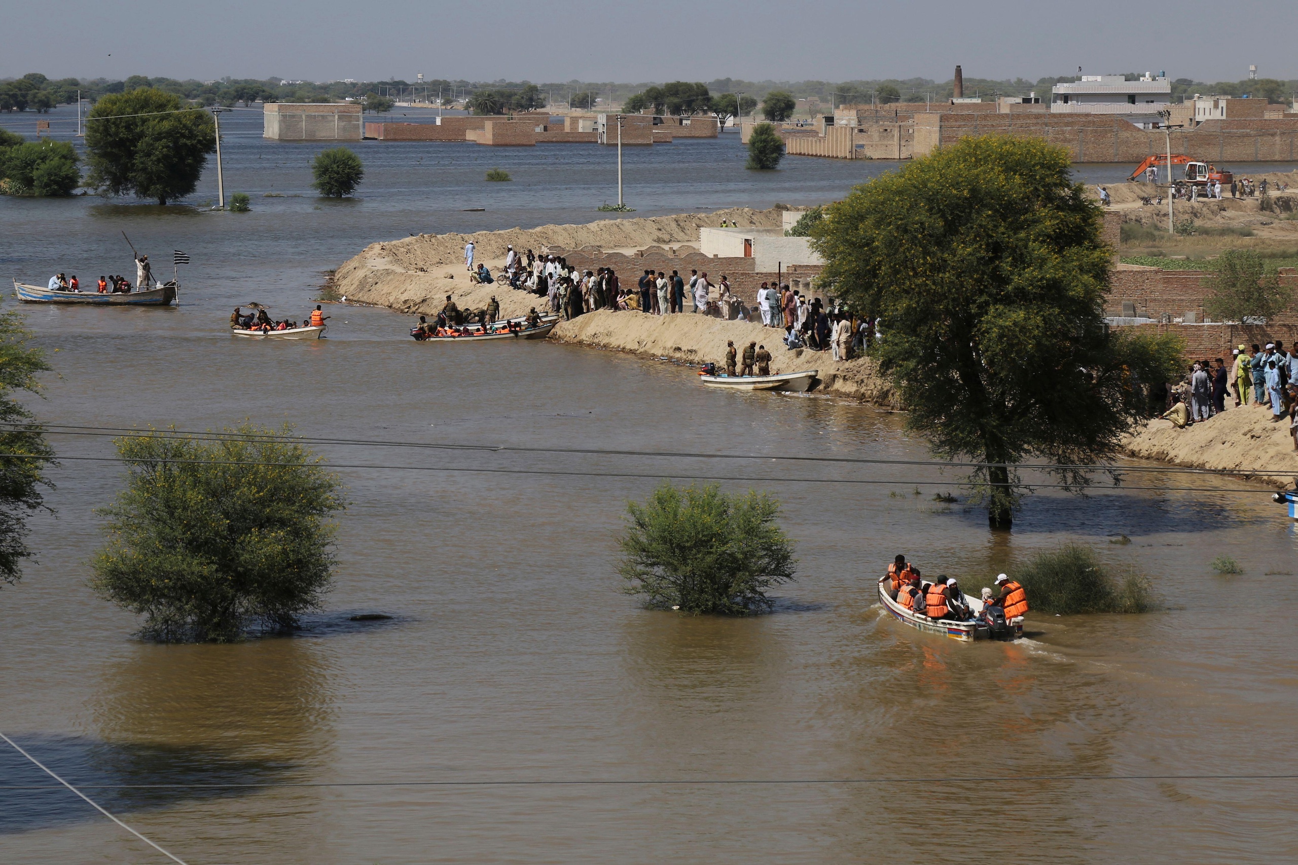 Rescue boats navigate floodwaters while people gather on the banks, amidst submerged buildings and trees in a flooded area.