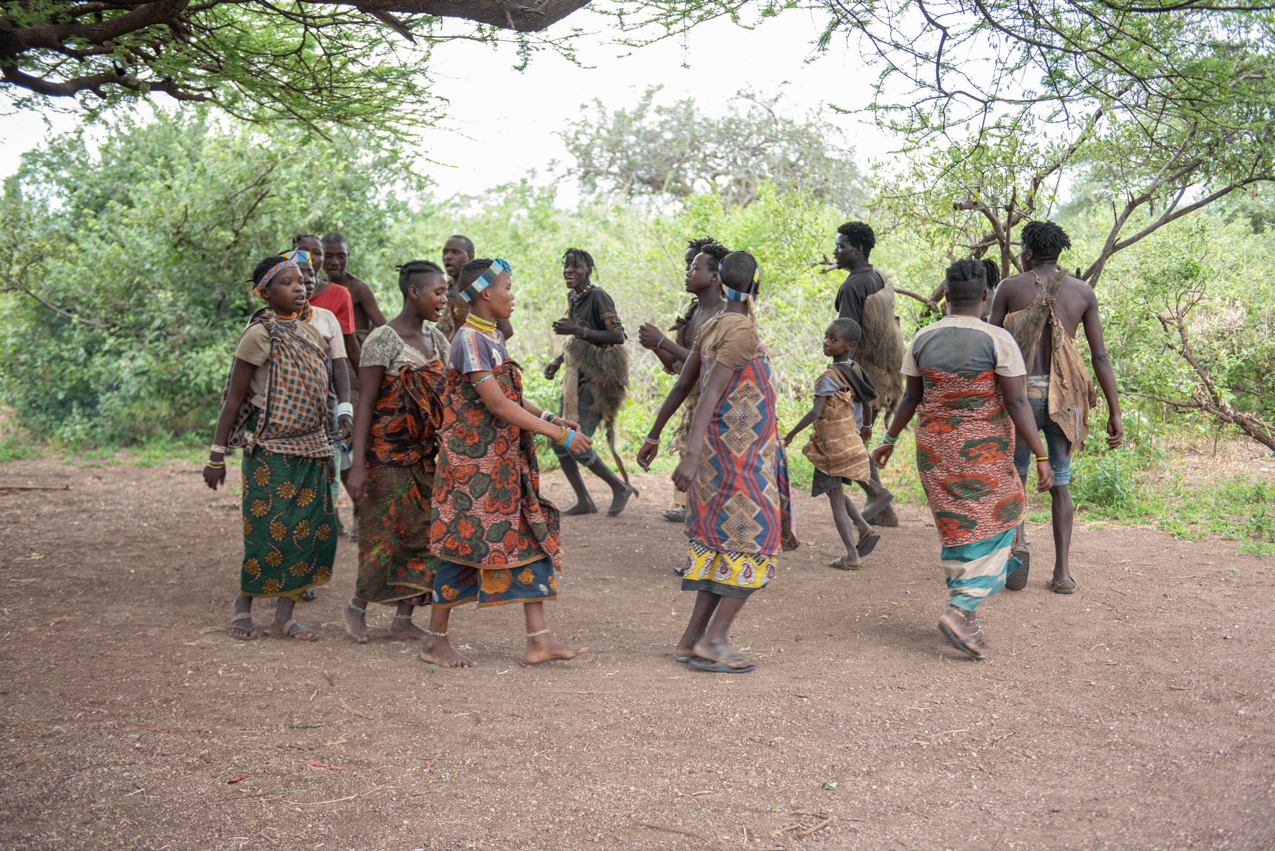 A group of people in colorful traditional clothing dance in a circle outdoors, under trees