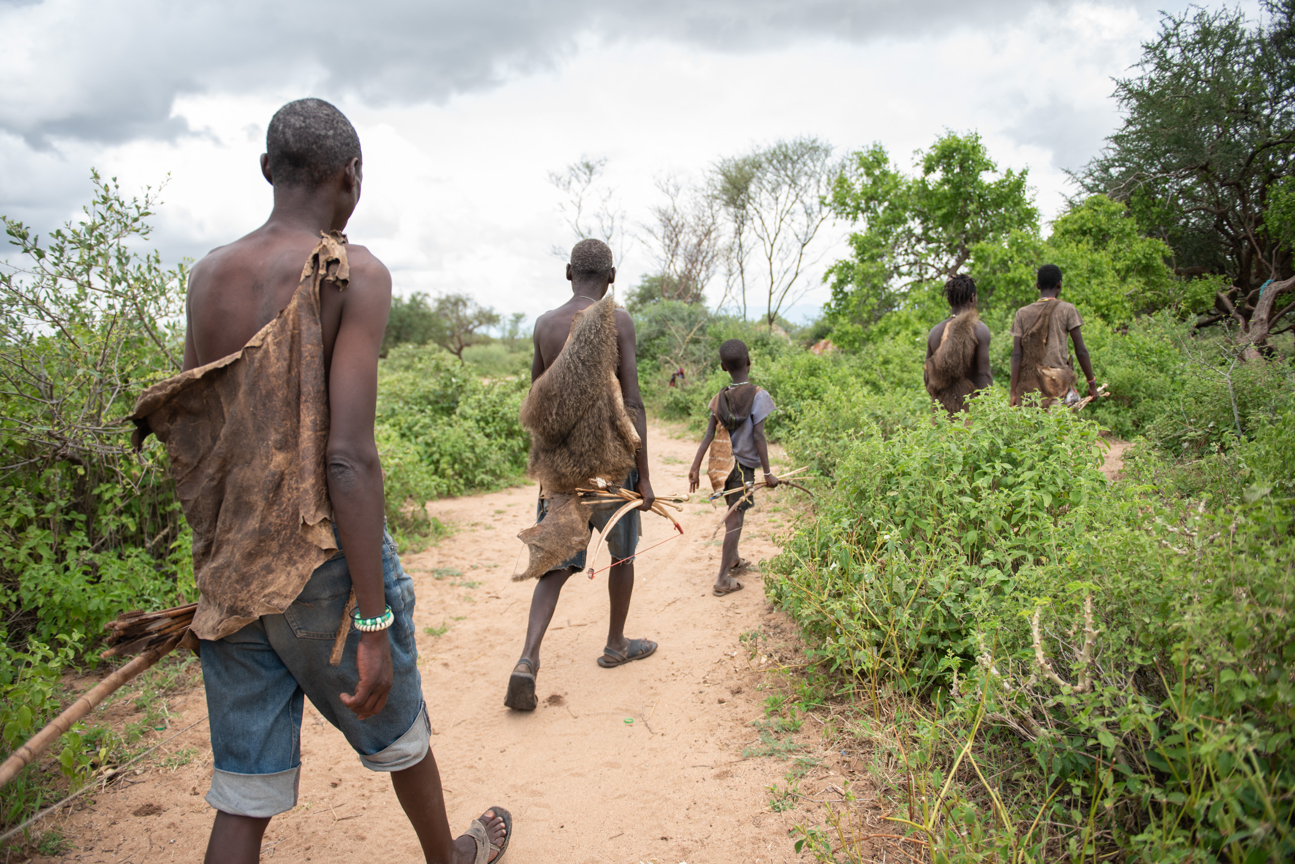 A group of young men wearing traditional clothing made from animal hides walk along a sandy path surrounded by greenery.
