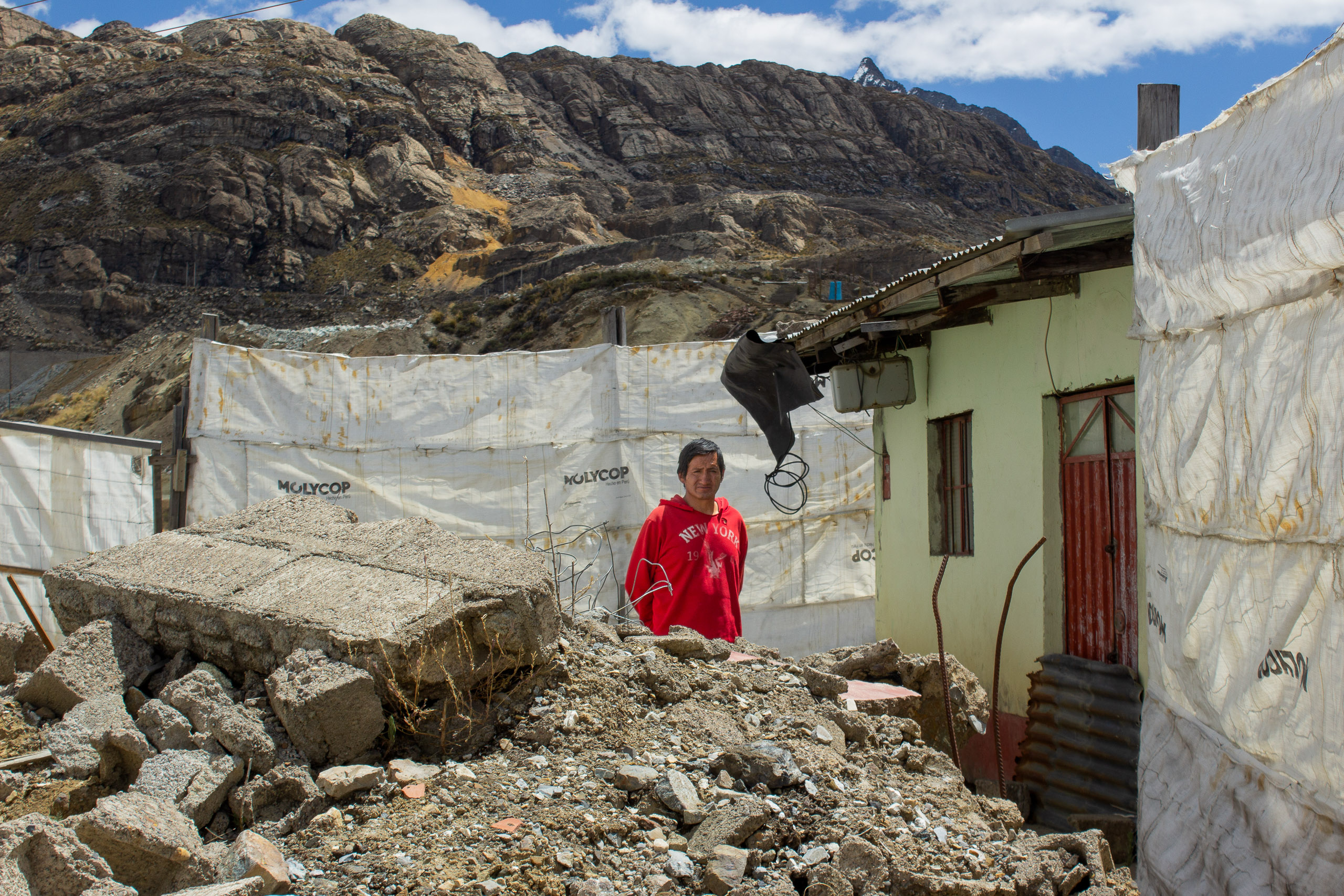 A person in a red shirt stands amid rubble and debris beside a worn building