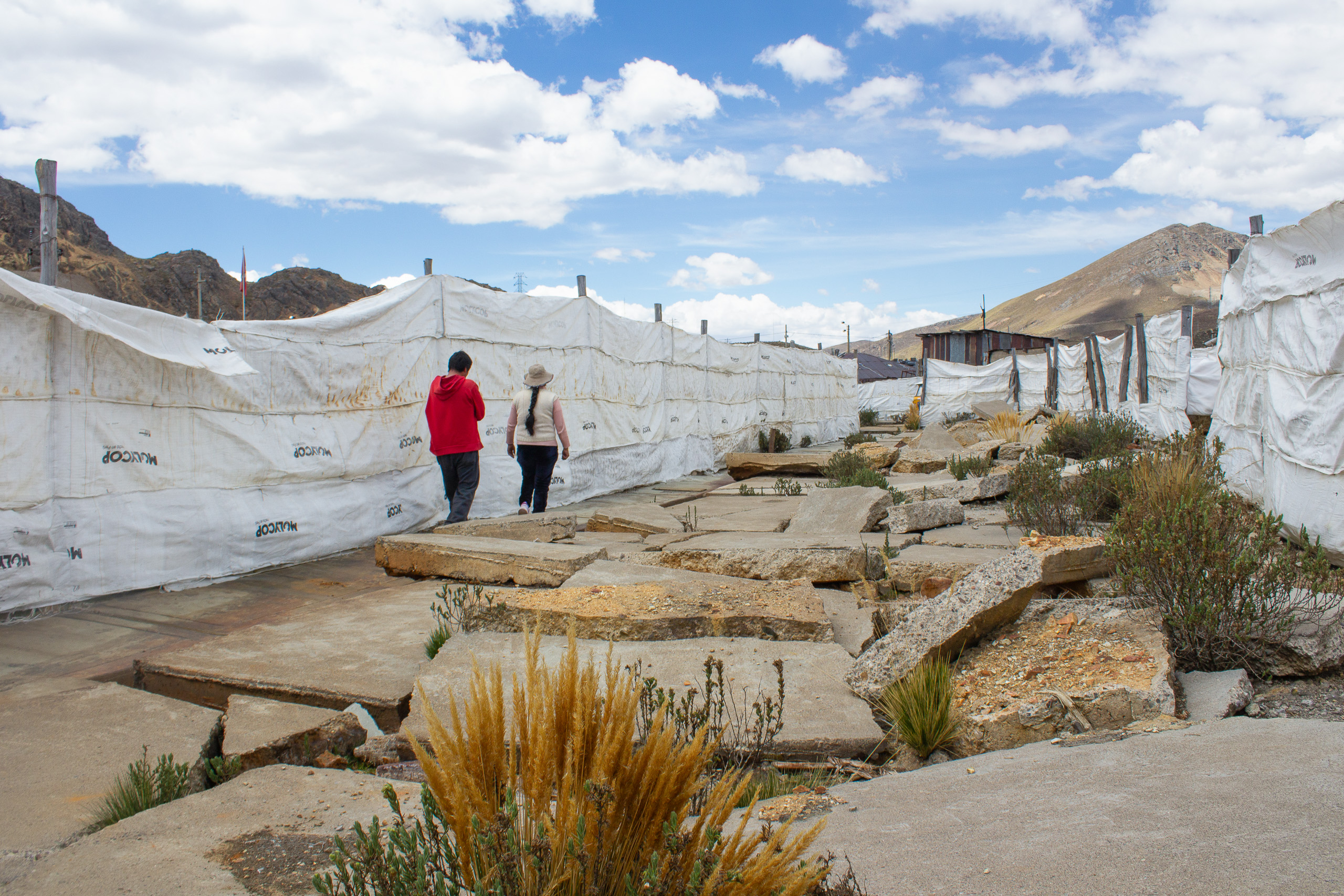 Two people walk along a path bordered by white tarps and rugged terrain