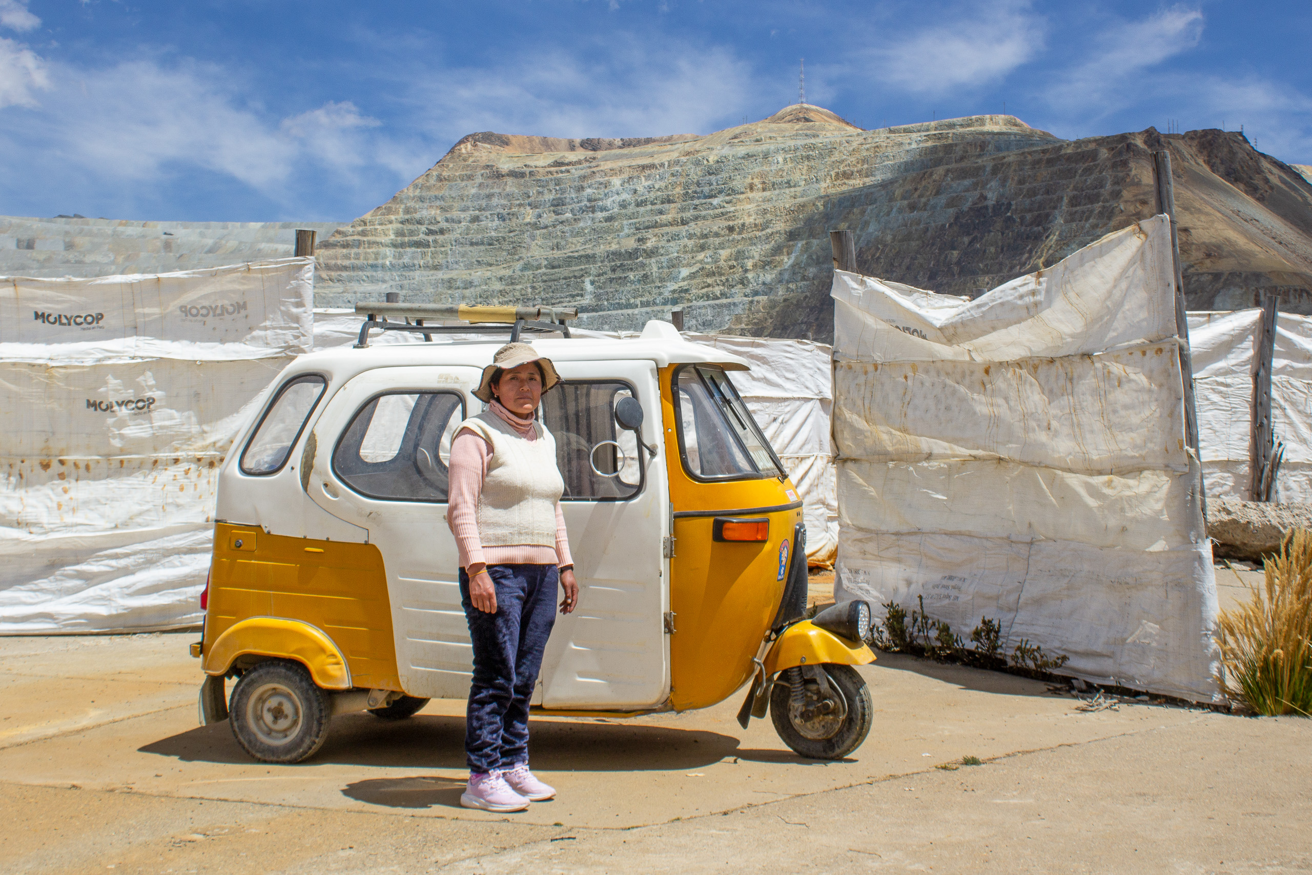 A woman stands next to a yellow and white tuk-tuk, in front of an opencast mine site
