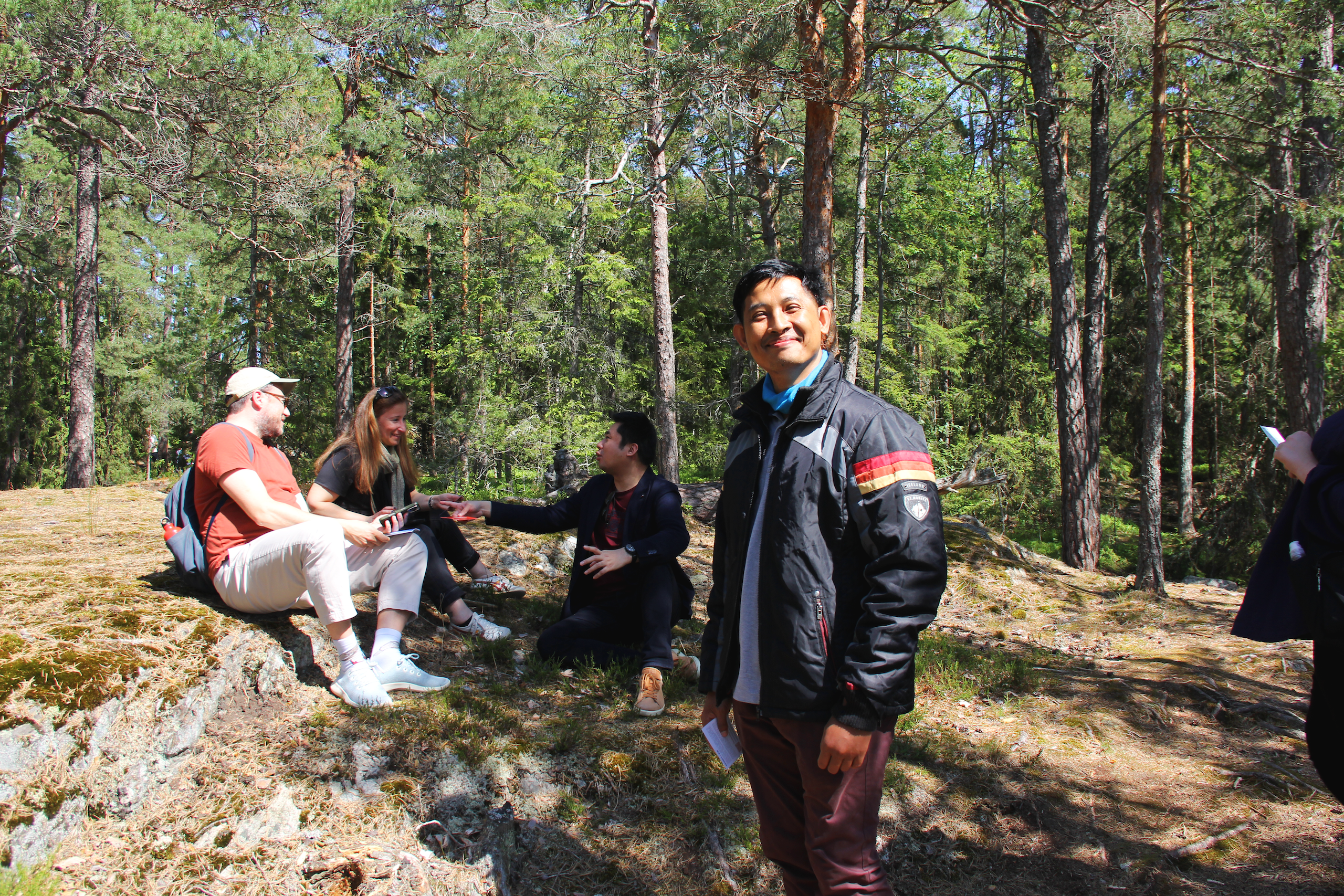A group of people engaged in discussion outdoors, surrounded by tall trees