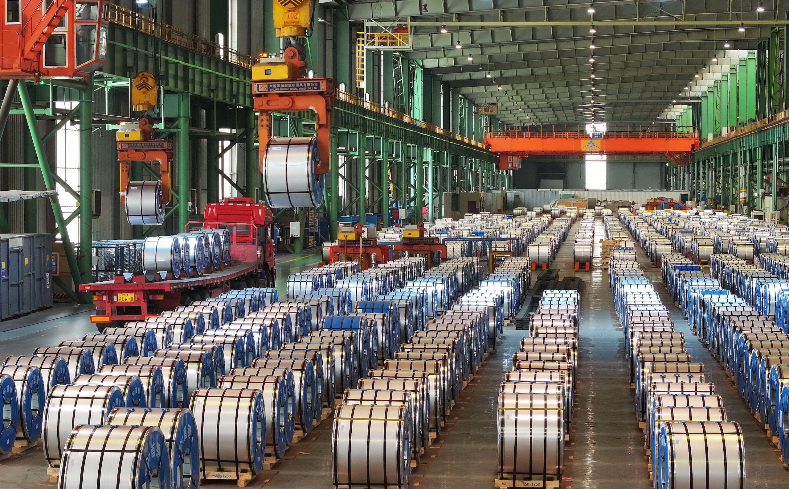 A warehouse filled with rows of steel coils on pallets, with cranes overhead lifting materials and a truck loading supplies.