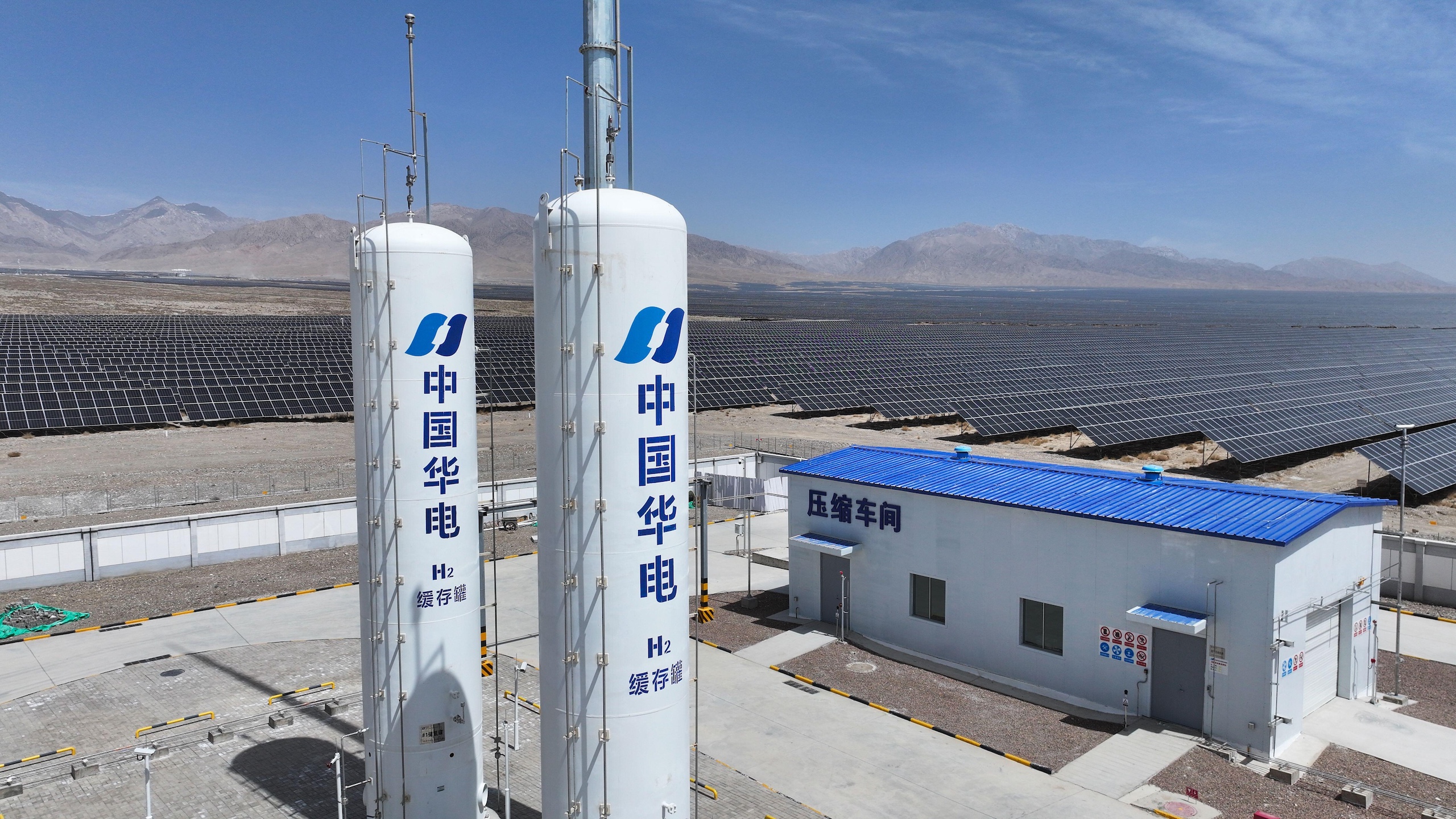 Two tall white hydrogen storage tanks with Chinese characters stand beside a blue-roofed building, set against a vast solar panel field and mountains.