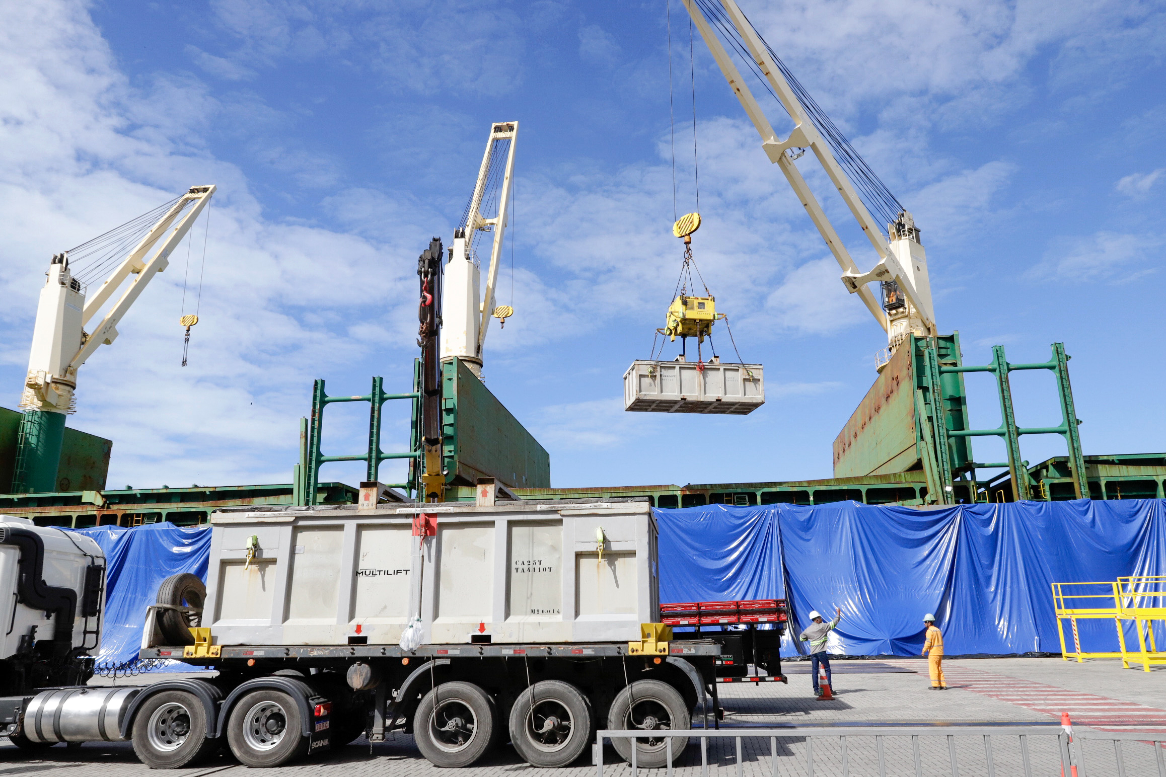 Two cranes load a container onto a truck at a port, with workers managing the operation in the background