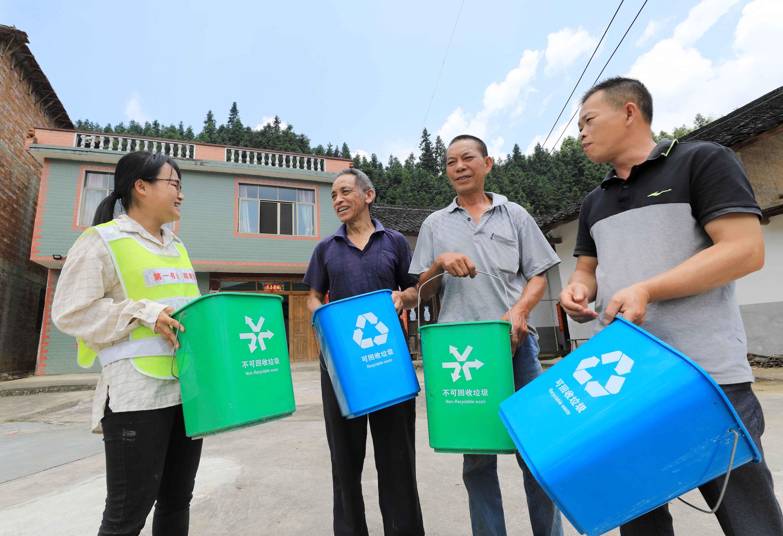 Four individuals hold recycling bins labeled for non-recyclable waste and recyclable materials, in front of a rural house surrounded by trees.
