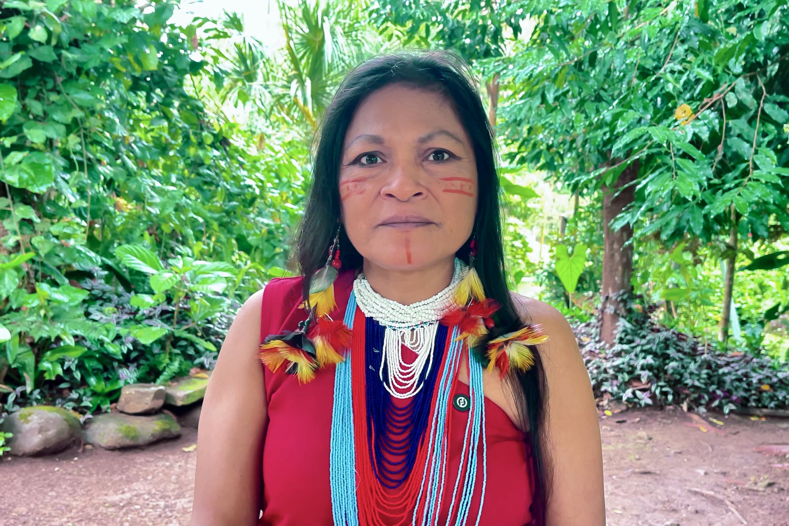 A woman adorned in vibrant traditional attire and colourful beadwork stands in a lush green outdoor area