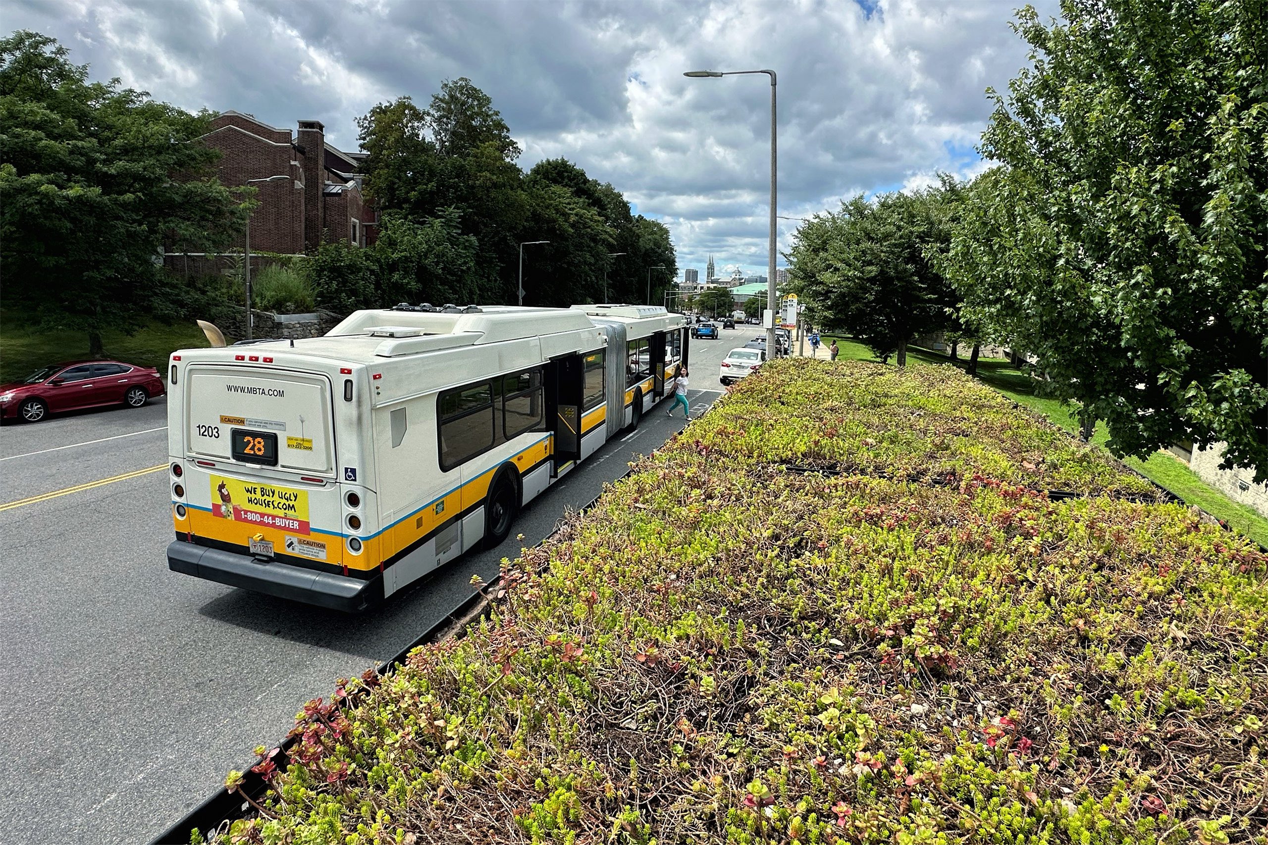 An air conditioned bus waits in traffic beside a landscaped area, with trees and greenery