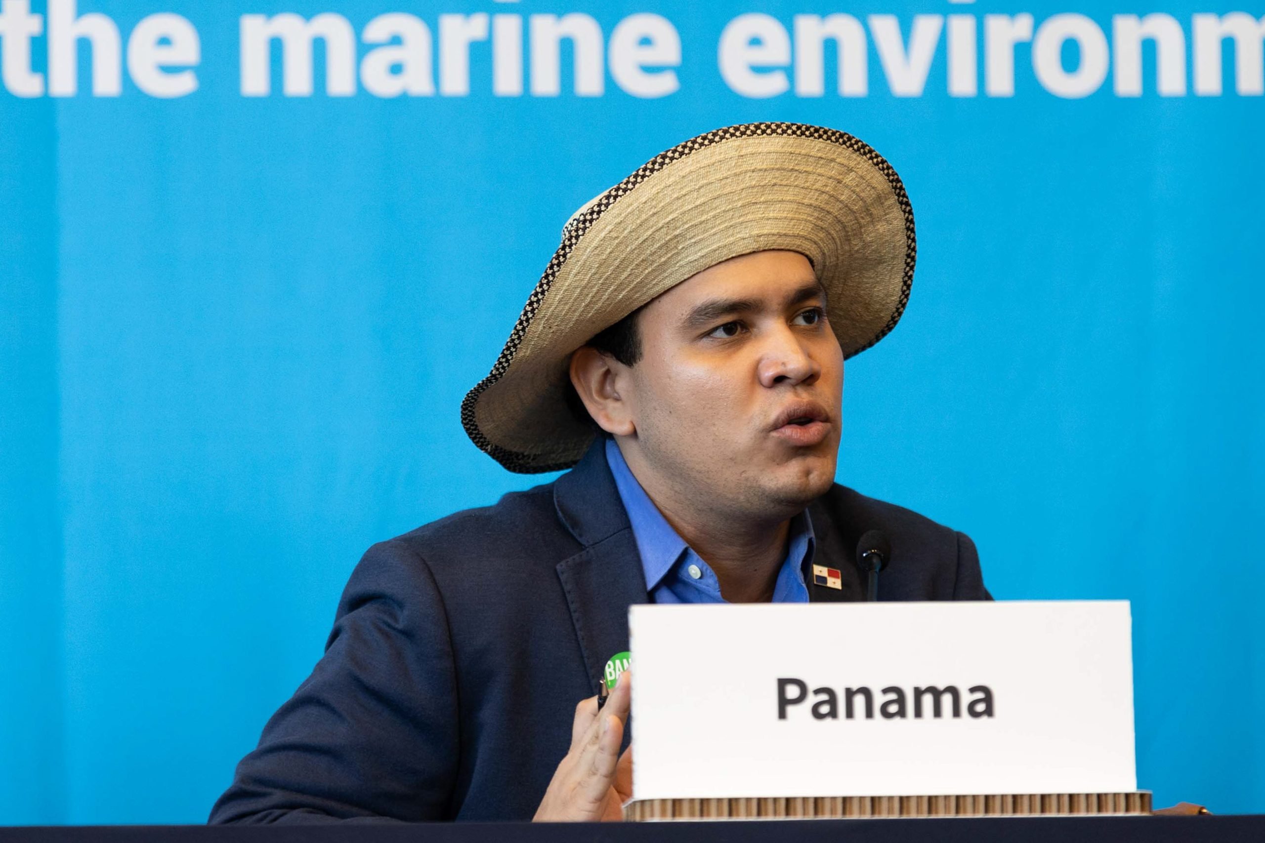 Una persona con sombrero y saco oscuro está sentada en una mesa de conferencias, con un letrero que dice "Panamá" al frente