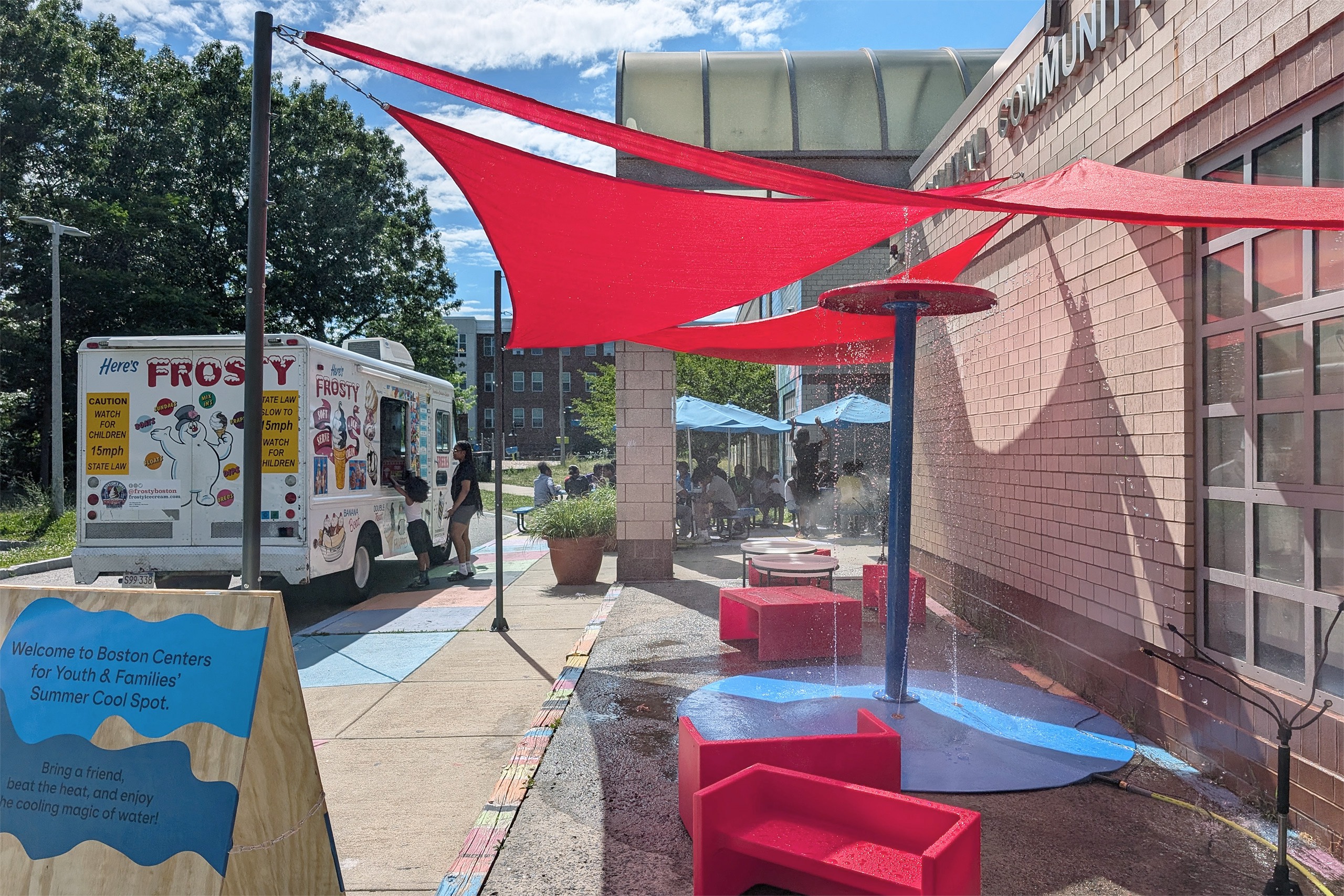 Asientos de colores bajo toldos rojos en un centro comunitario, junto a un camión de helados