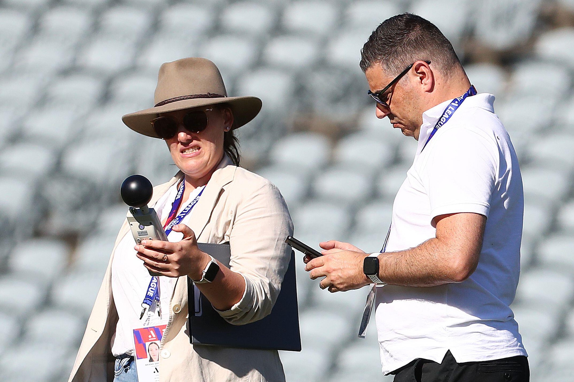 two match officials use a Wet Bulb Globe Temperature device to measure the temperature in a stadium