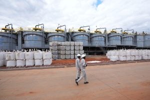 Two workers in safety gear walk past large storage tanks and stacked containers in a plant site