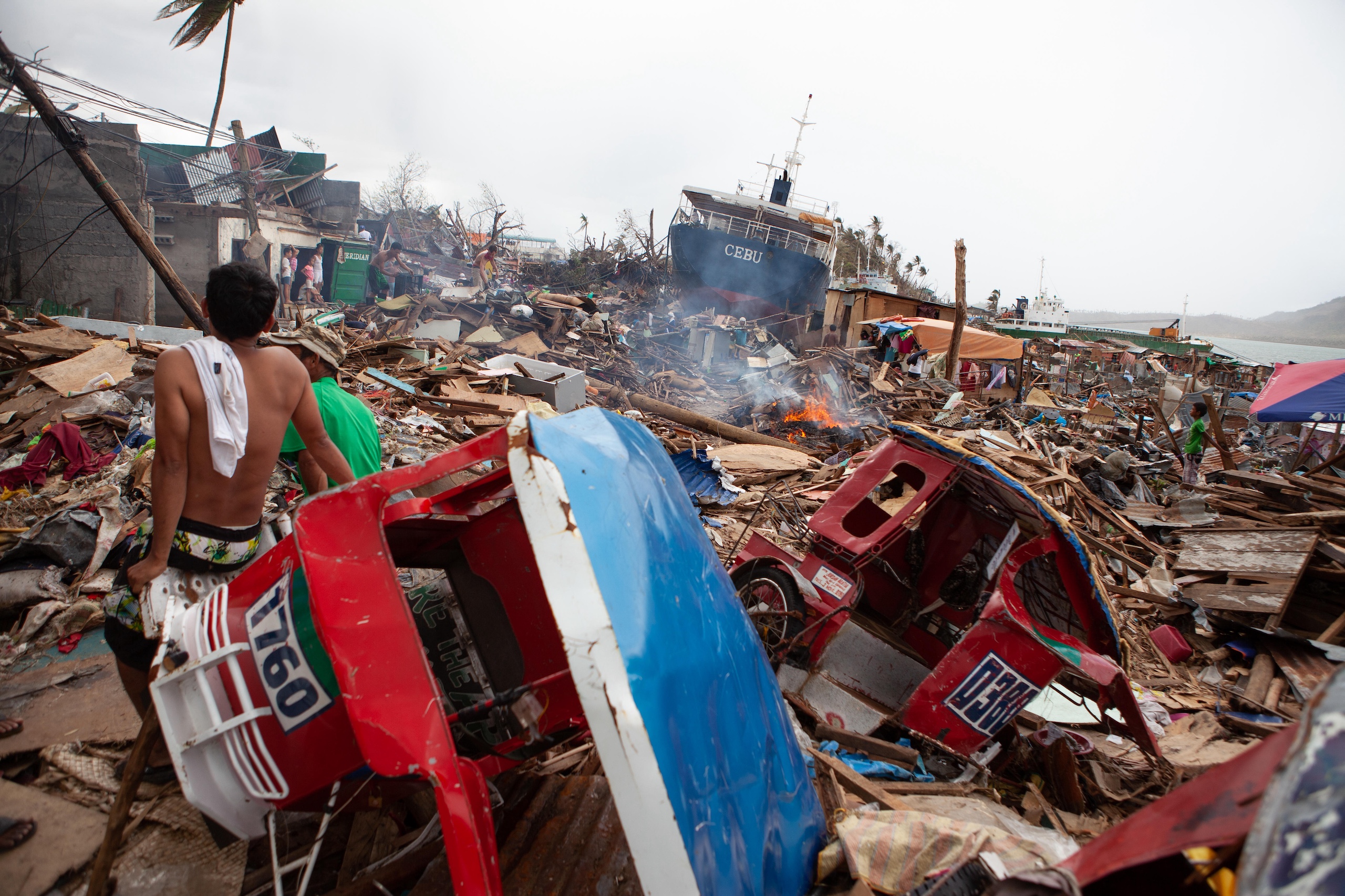 Devastated landscape post-disaster, featuring destroyed homes, debris, overturned vehicles, and a ship in the background